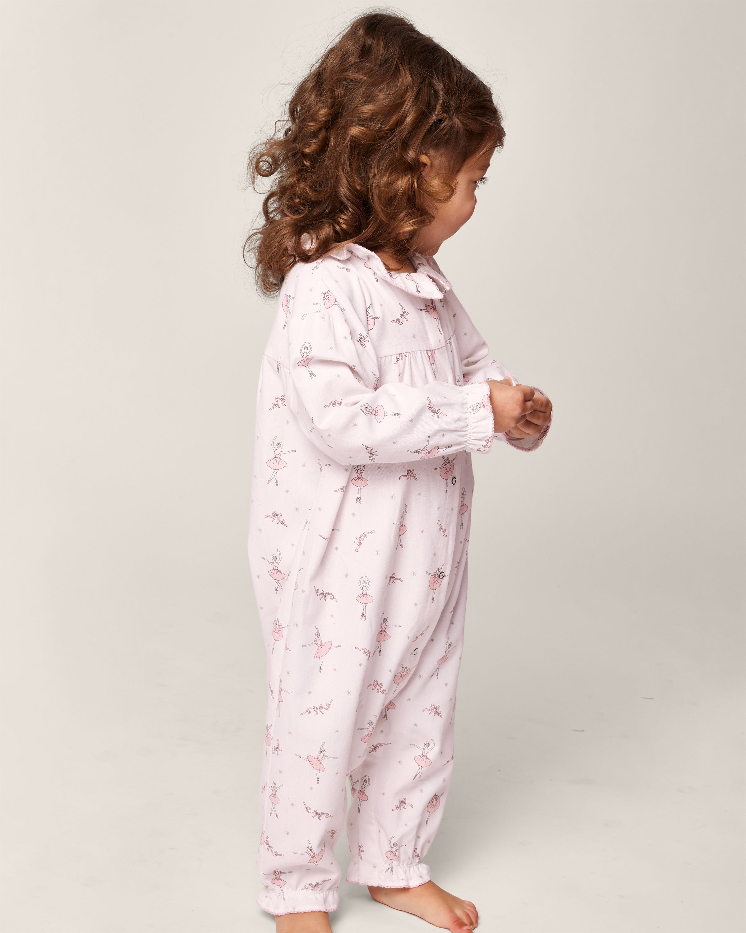 A young child with curly hair stands sideways wearing Petite Plumes Babys Flannel Madeline Romper in Sugar Plum Fairy print. Made from flame-retardant fabric, this stylish childrens sleepwear ensures safety against a plain background.