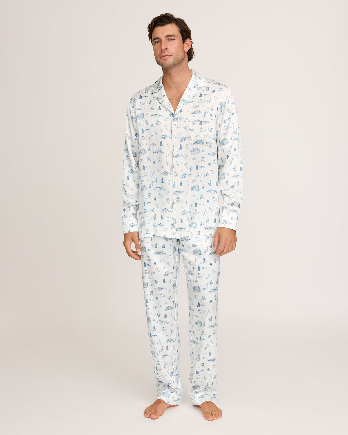 A man stands barefoot against a plain white background, wearing the Petite Plume x White Elephant Men’s Silk Pajama Set—a matching ensemble of white pajama pants and a long-sleeve button-up shirt featuring a subtle light blue pattern.