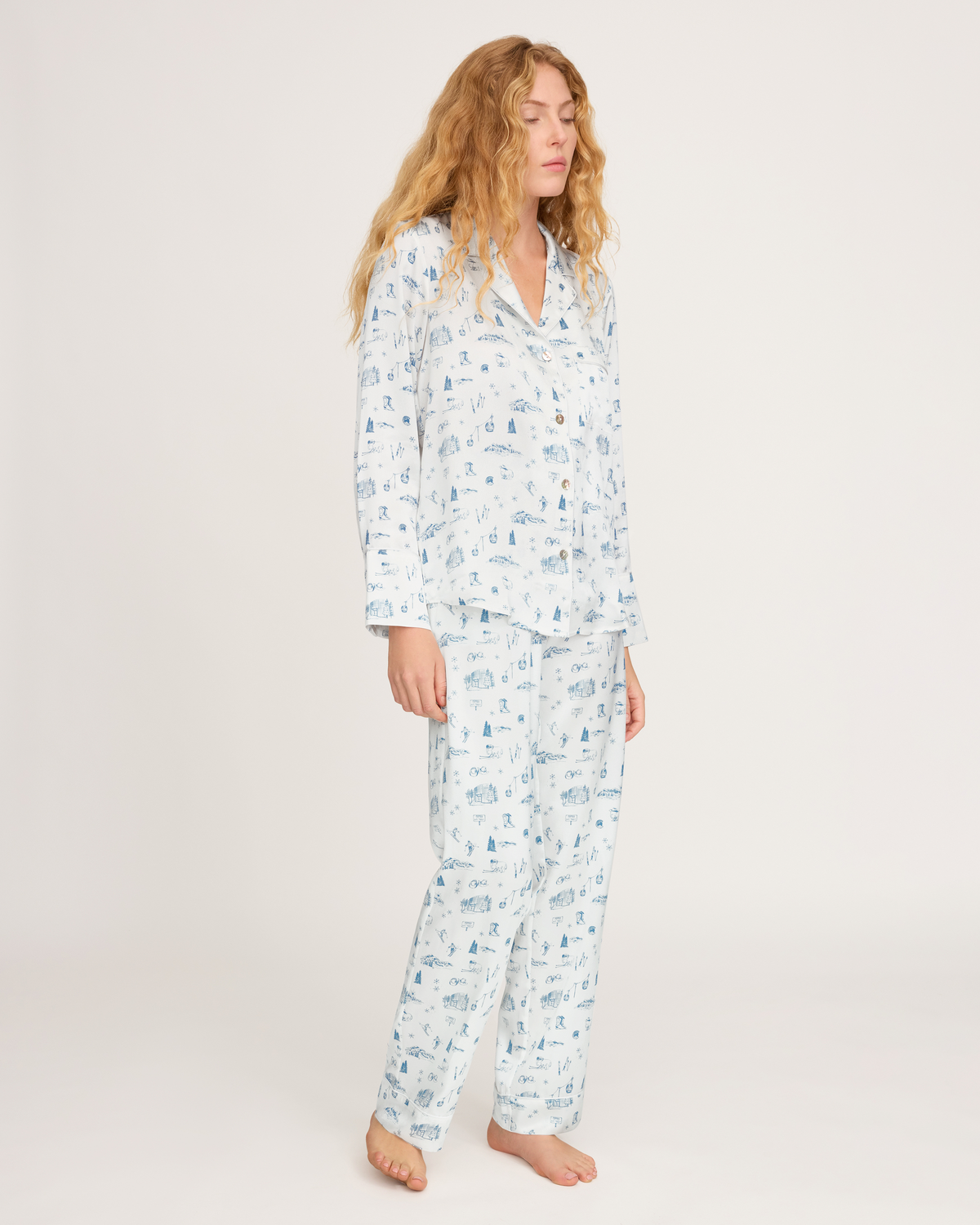 A woman with long wavy hair stands barefoot against a plain white background, wearing the Petite Plume x White Elephant Women’s Silk Pajama Set—a light blue and white printed pajama ensemble featuring a button-up top and matching pants.