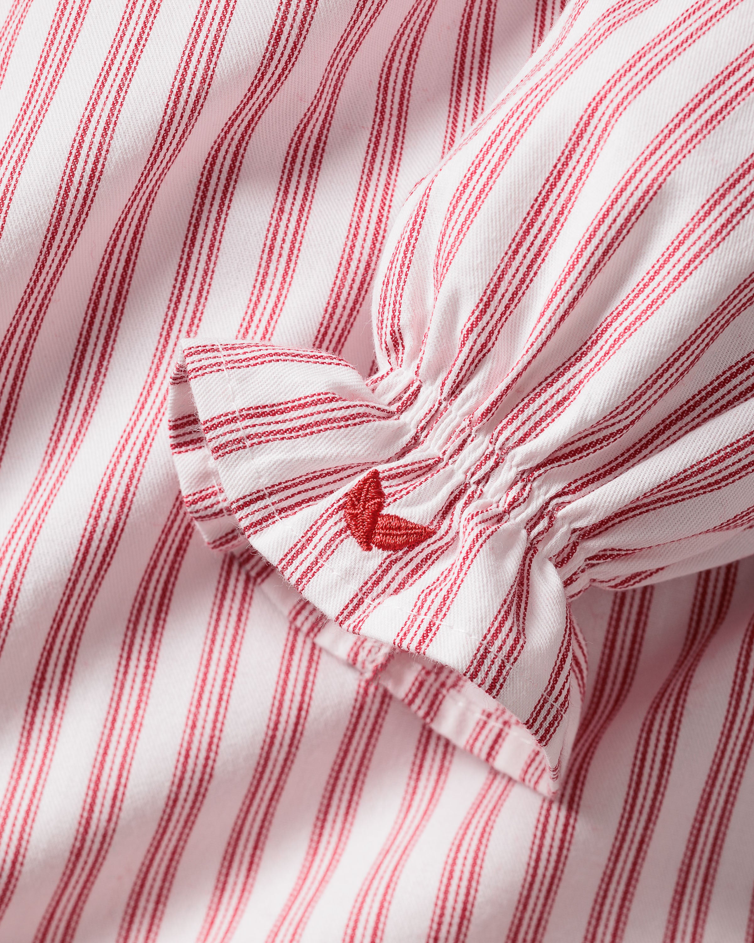 Close-up of Petite Plumes Girls Twill Delphine Nightgown in Antique Red Ticking, featuring pink and white stripes with red stitched ruffles and an embroidered star. Ideal for childrens sleepwear, its smooth texture highlights the detailed pattern and craftsmanship.