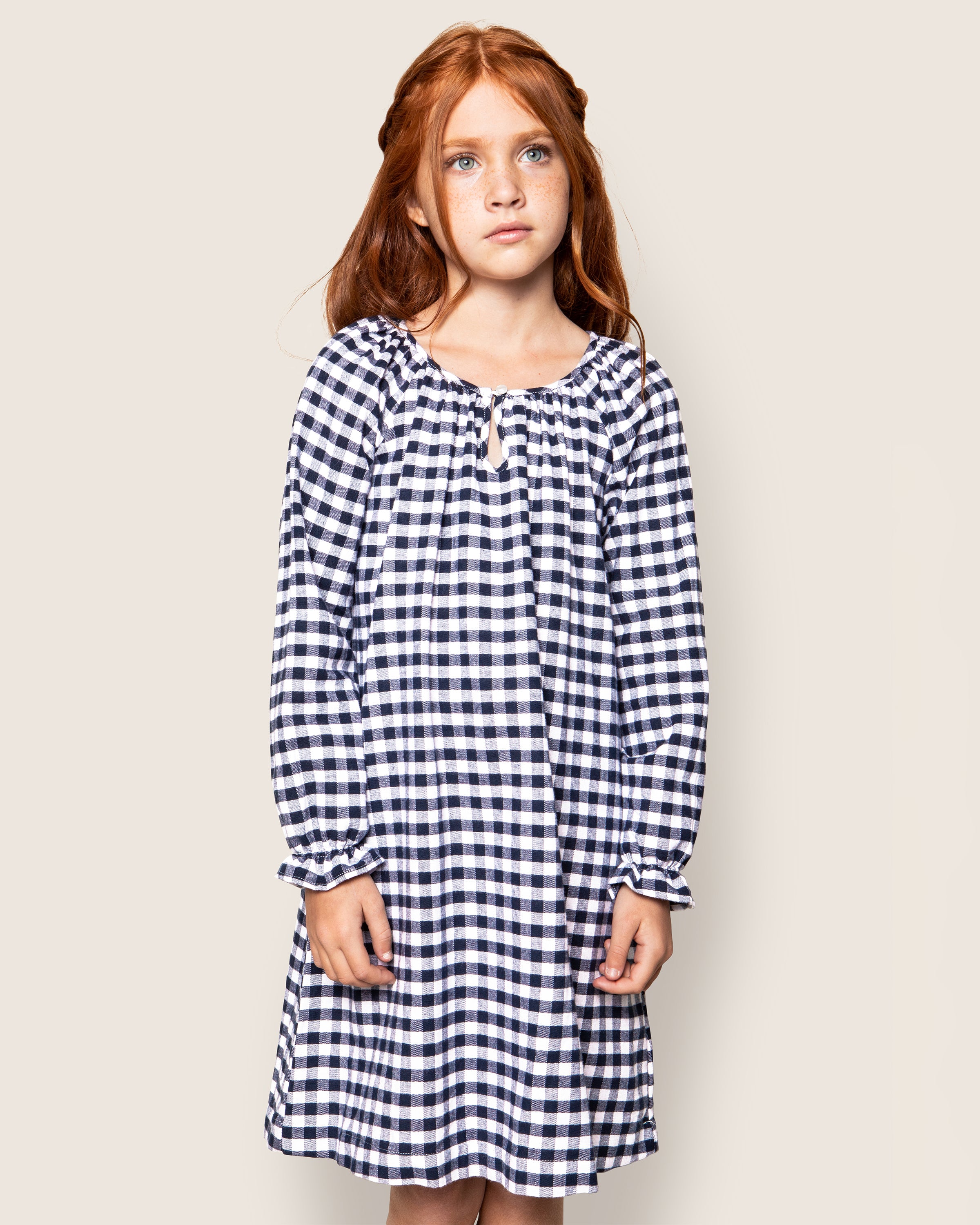 A girl with long red hair stands against a plain background, wearing the Girls Flannel Delphine Nightgown in Navy Gingham by Petite Plume. She looks forward with a neutral expression, highlighting the classic style.