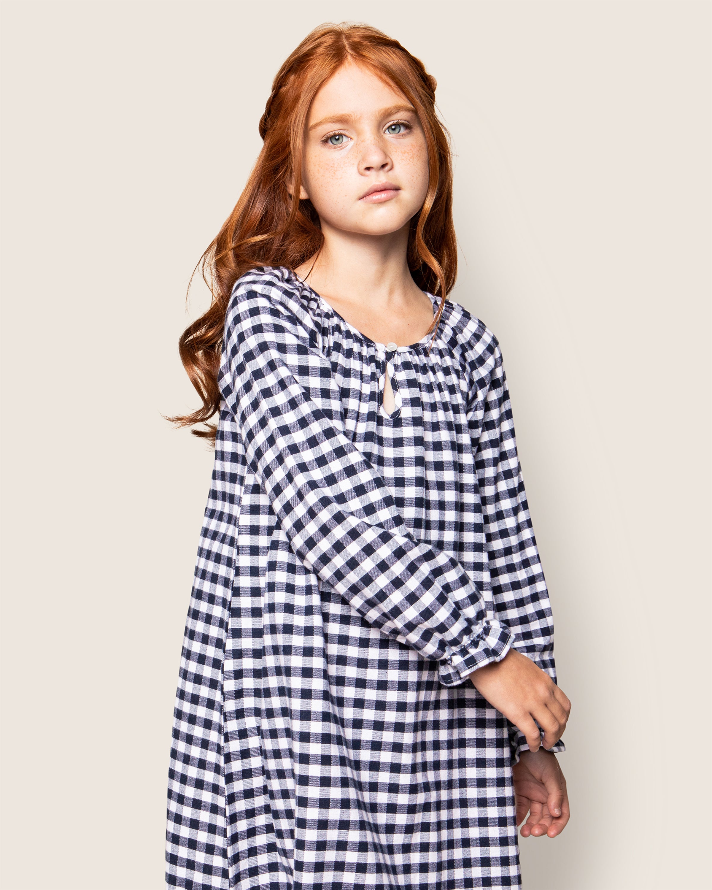 A young girl with long red hair, wearing a Petite Plume Girls Flannel Delphine Nightgown in Navy Gingham, stands against a plain background. Her calm expression is complemented by her relaxed arms hanging gently by her side.