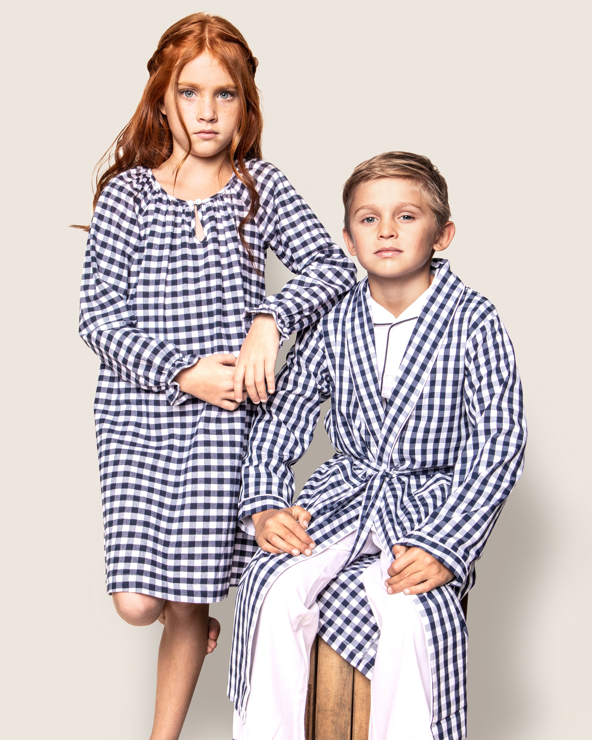 Two children wearing matching gingham pajamas pose against a plain background. One wears a Petite Plume Girls Flannel Delphine Nightgown in Navy Gingham, while the other wears a robe over flannel sleepwear, sitting on a stool. Their expressions are calm and serene.