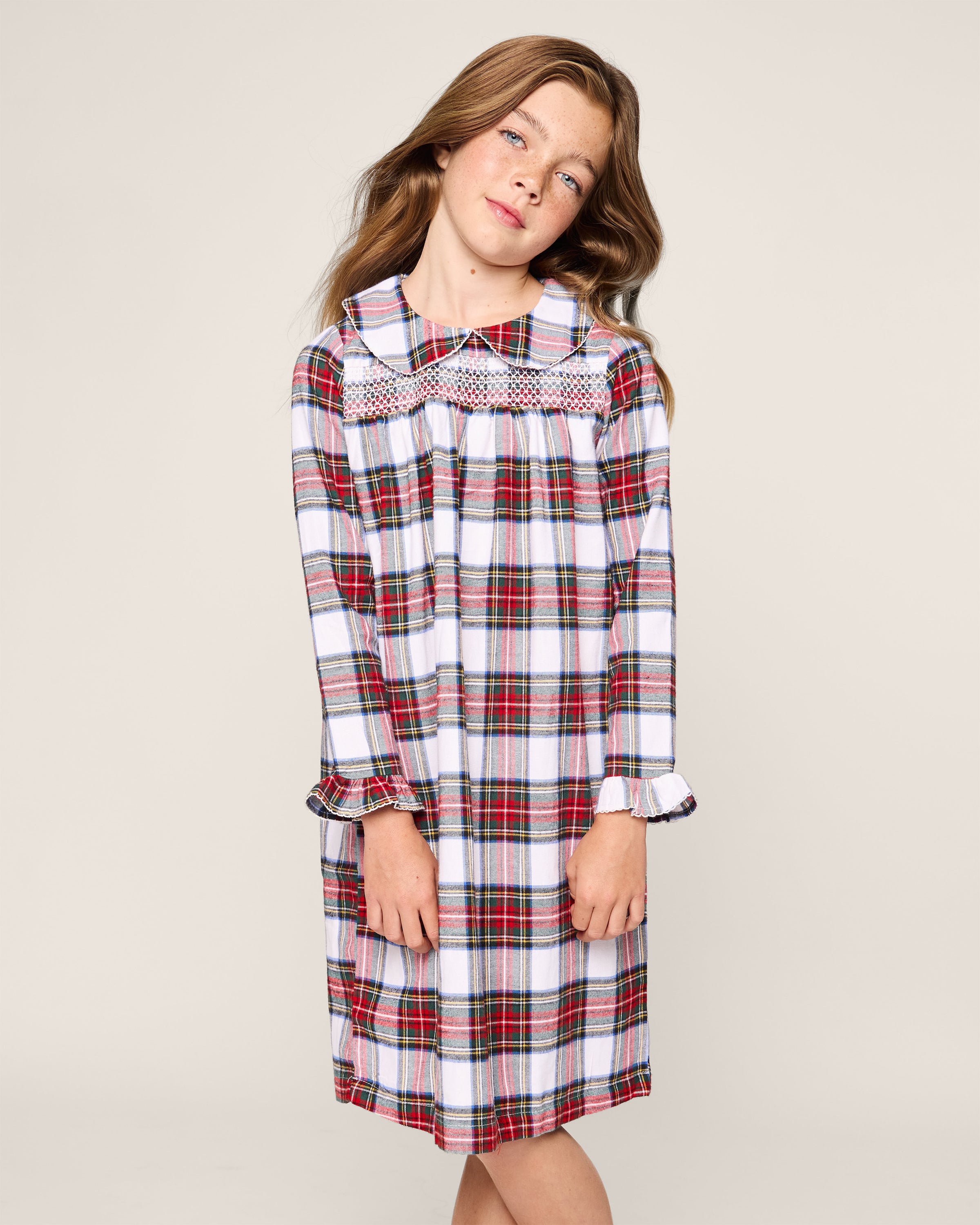 A young girl in Petite Plumes Brushed Cotton Eloise Nightgown, featuring Balmoral Tartan and a white collar, stands against a plain background looking slightly to the side. Her hands are clasped in front of her, and her hair cascades over her shoulders.