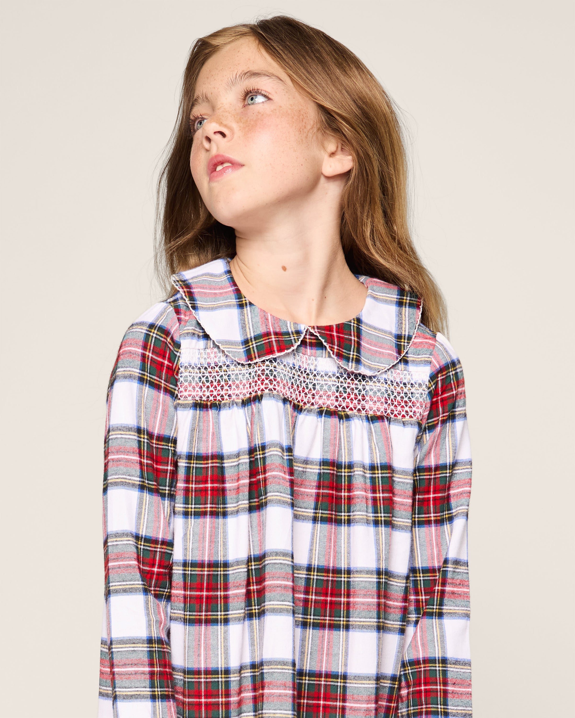 A young girl with long brown hair gazes sideways, donning the Petite Plume Girls Brushed Cotton Eloise Nightgown in Balmoral Tartan. It features red, blue, and white yarn-dyed stripes and a smocked chest with a white Peter Pan collar. The backdrop is plain and light-colored.