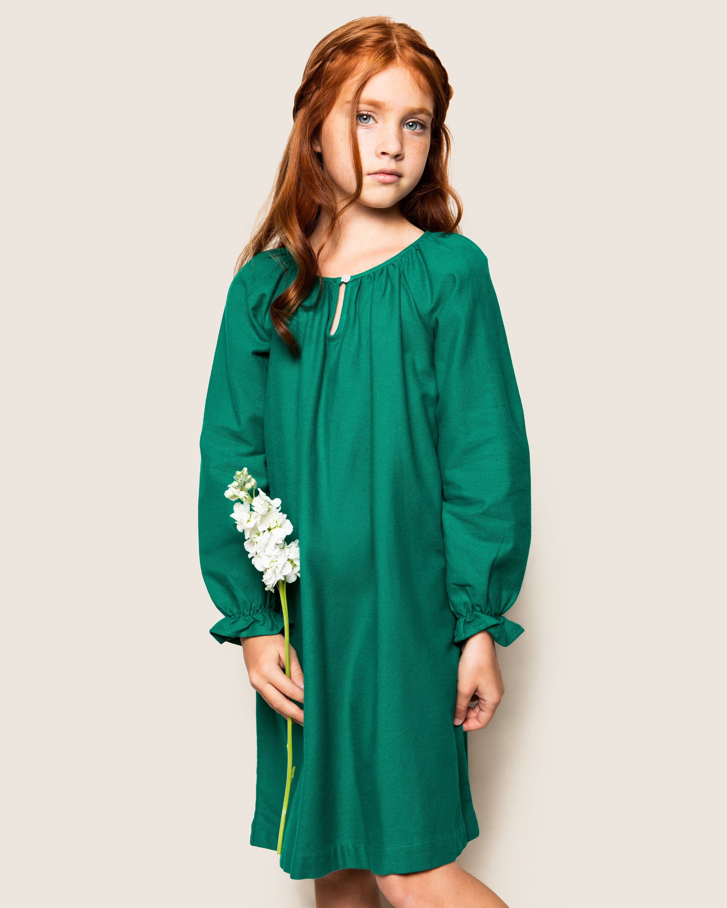 A young girl with long red hair stands against a plain background, wearing Petite Plumes Girls Flannel Delphine Nightgown in Green. She holds a single white flower in one hand, gazing forward with a neutral expression.