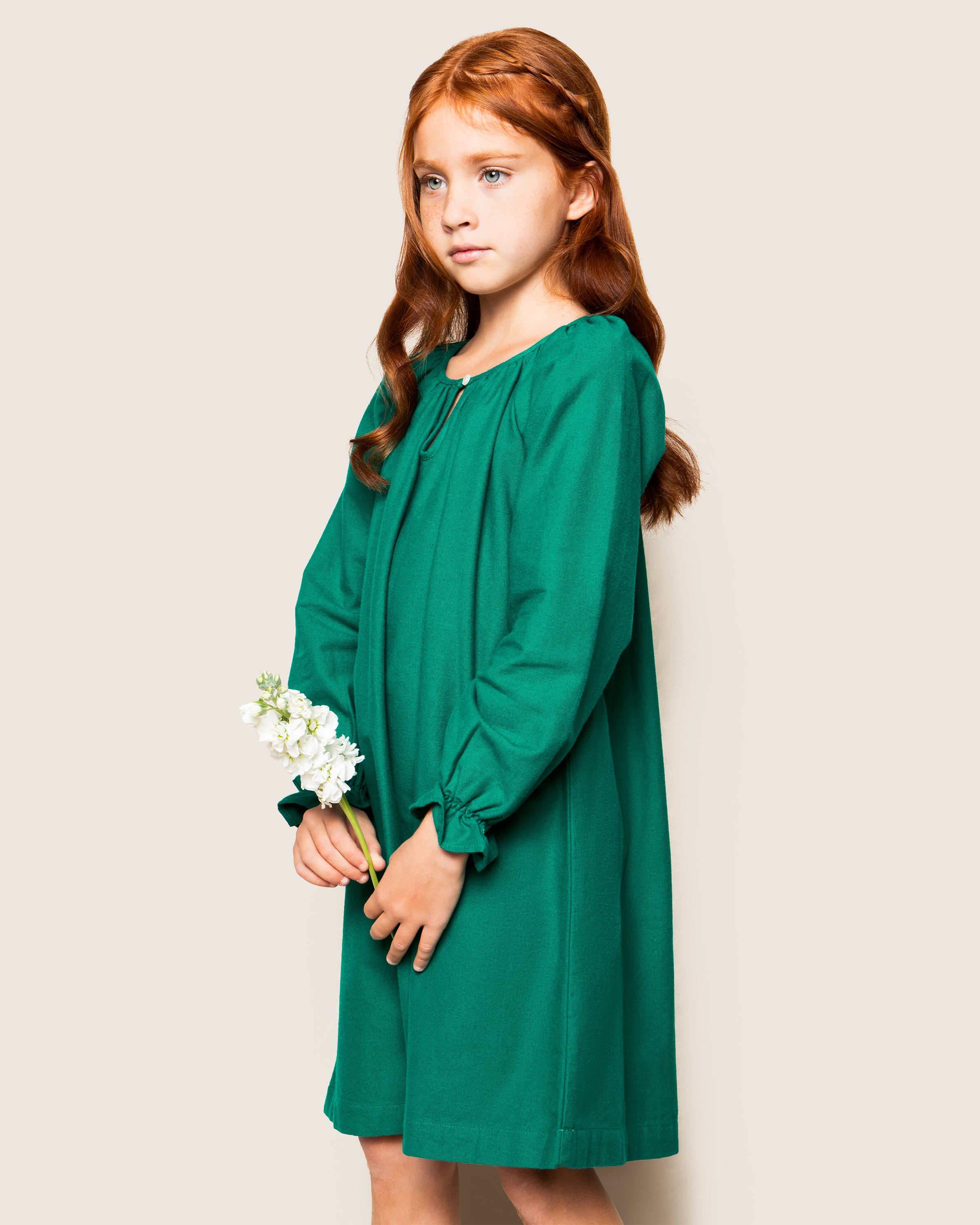 A young girl with long red hair holds white flowers while wearing Petite Plumes Girls Flannel Delphine Nightgown in Green against an off-white backdrop, gazing to the side.