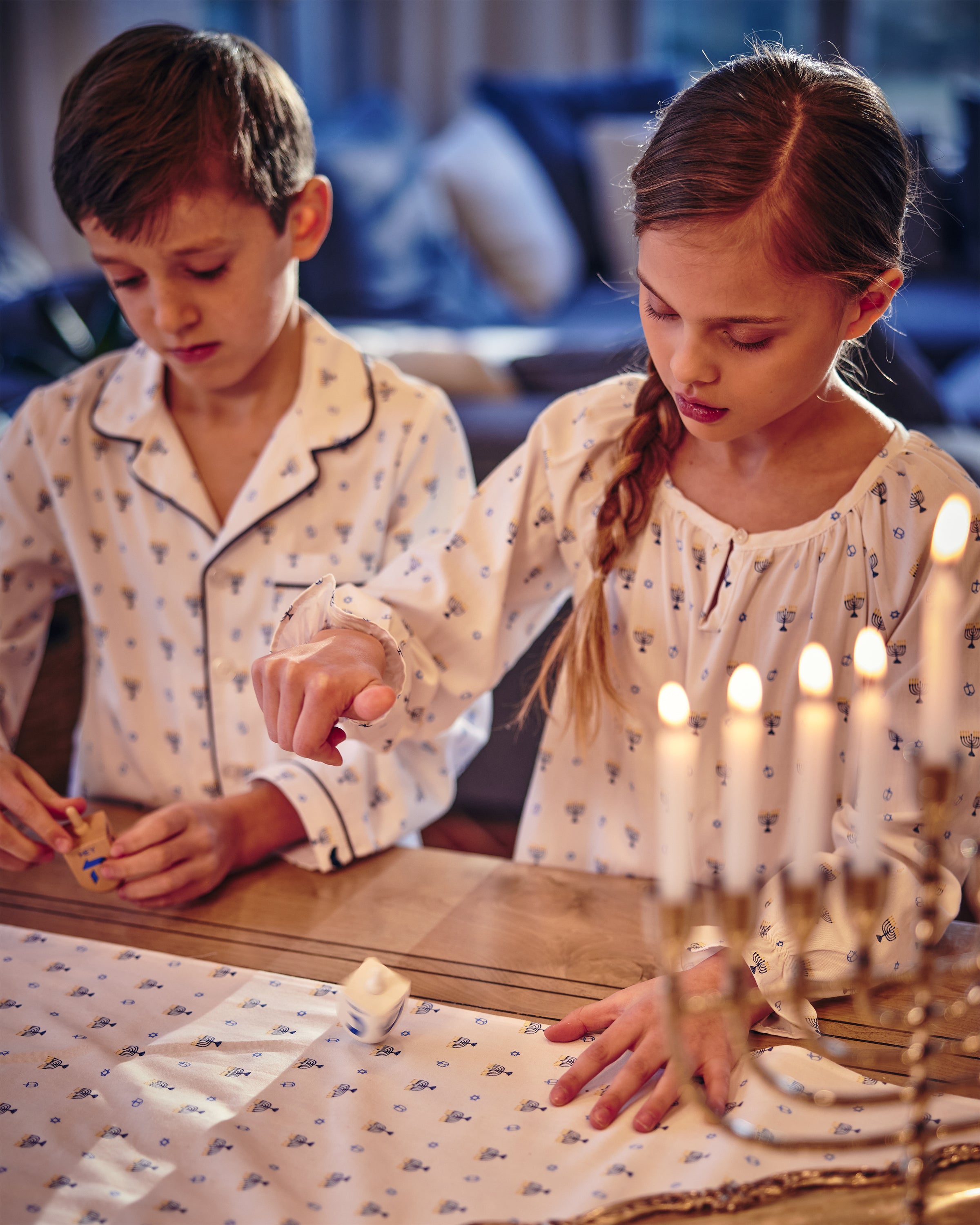 A young boy and girl, dressed in matching Petite Plume sleepwear, play dreidel on a wooden table. A lit menorah warmly glows nearby, highlighting the festive Hanukkah vibe and the coziness of their softly lit evening in a Girls Twill Delphine Nightgown in Happy Hanukkah.