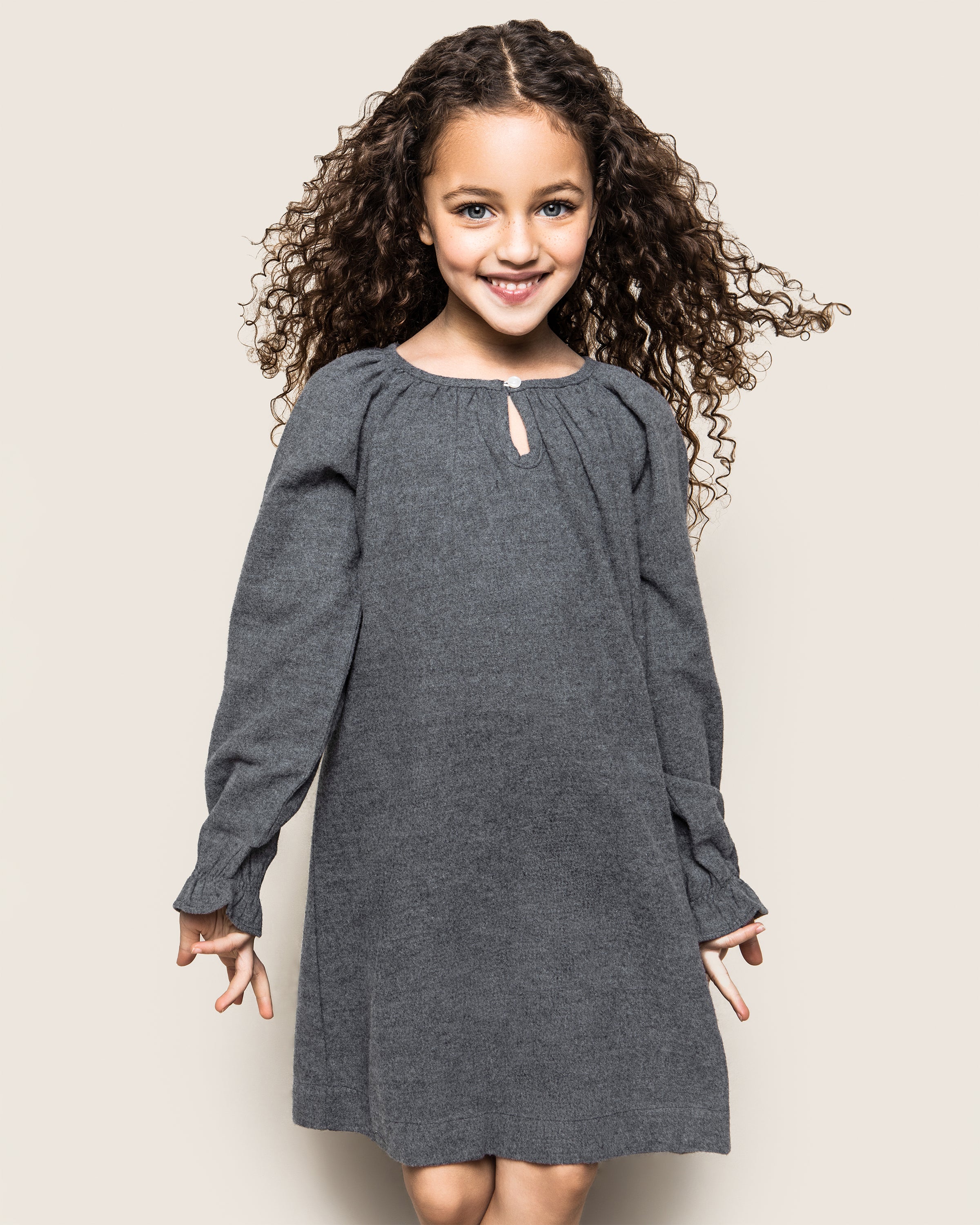 A curly-haired young girl smiles against a plain background, wearing Petite Plumes Girls Flannel Delphine Nightgown in Grey, her hands by her sides.