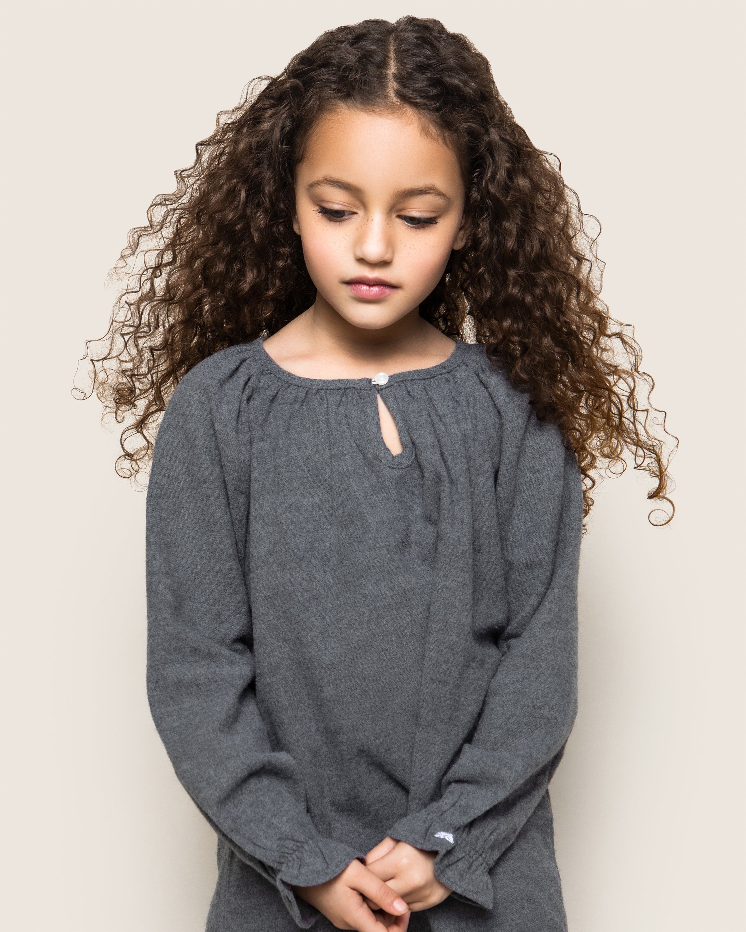 A young girl with long, curly brown hair is looking down, wearing the cozy Girls Flannel Delphine Nightgown in Grey by Petite Plume. The plain light beige background highlights the simplicity and comfort of her brushed cotton flannel attire.