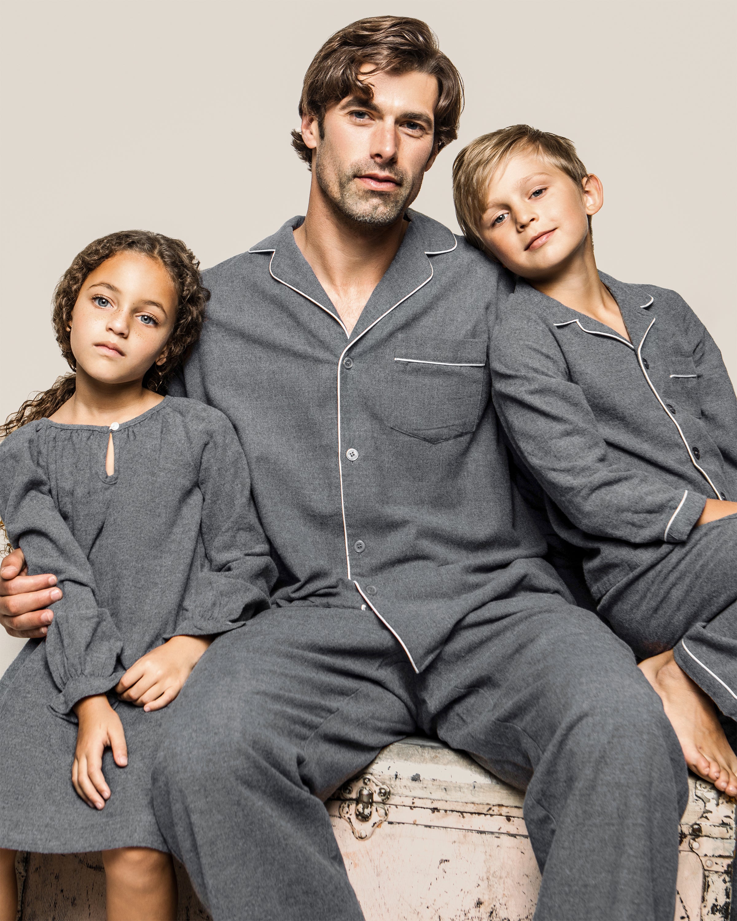 A man in brushed cotton flannel pajamas sits with a girl and boy, both in cozy gray outfits. The girl wears Petite Plumes Girls Flannel Delphine Nightgown in Grey. Their calm presence against the beige backdrop highlights their choice of chemical-free sleepwear.