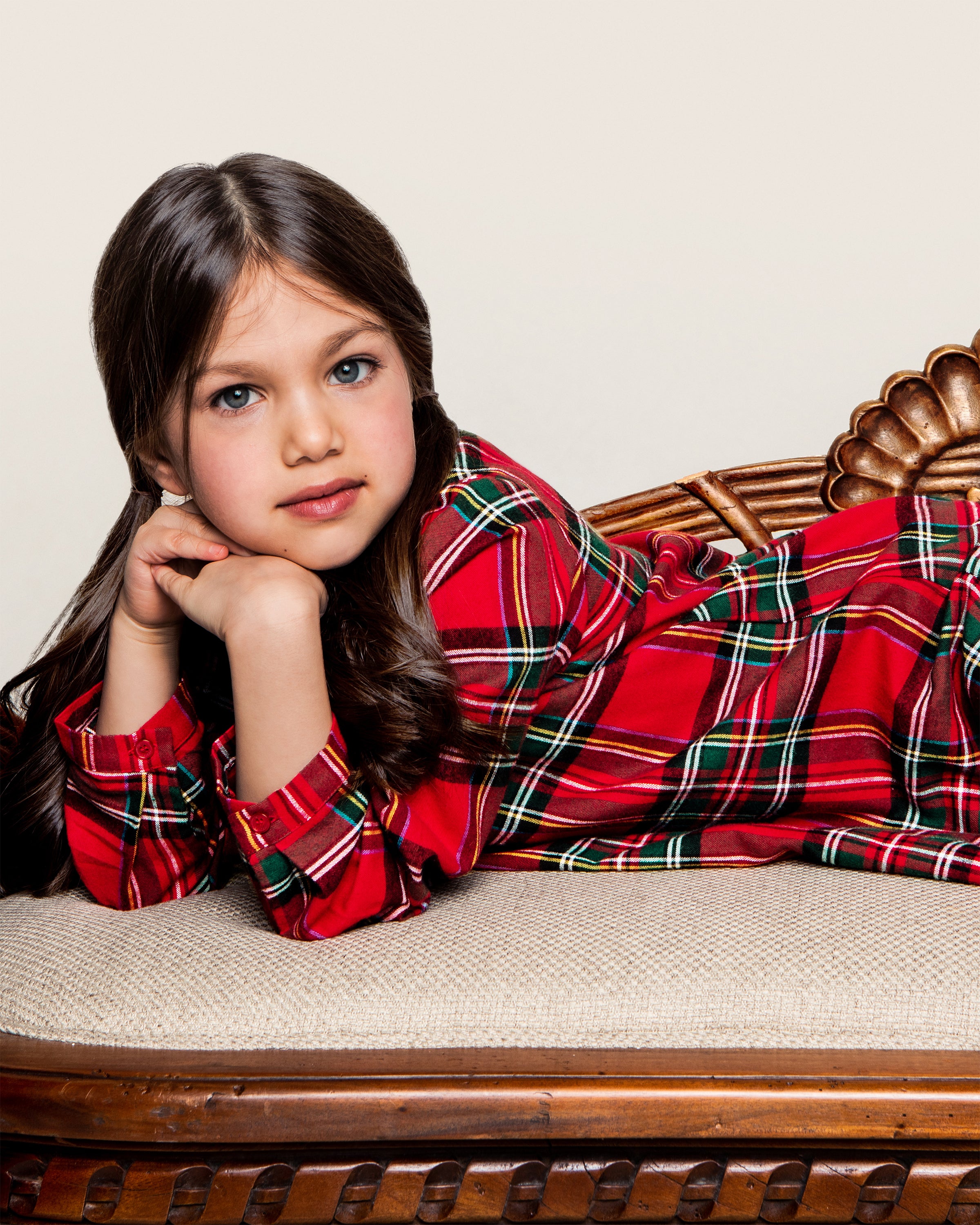 A young girl with long dark hair, wearing a Petite Plume Imperial Tartan Beatrice Nightgown, lies on her stomach on a cushioned wooden bench. She rests her chin on her hand and gazes at the camera with a relaxed expression against a plain light-colored background.