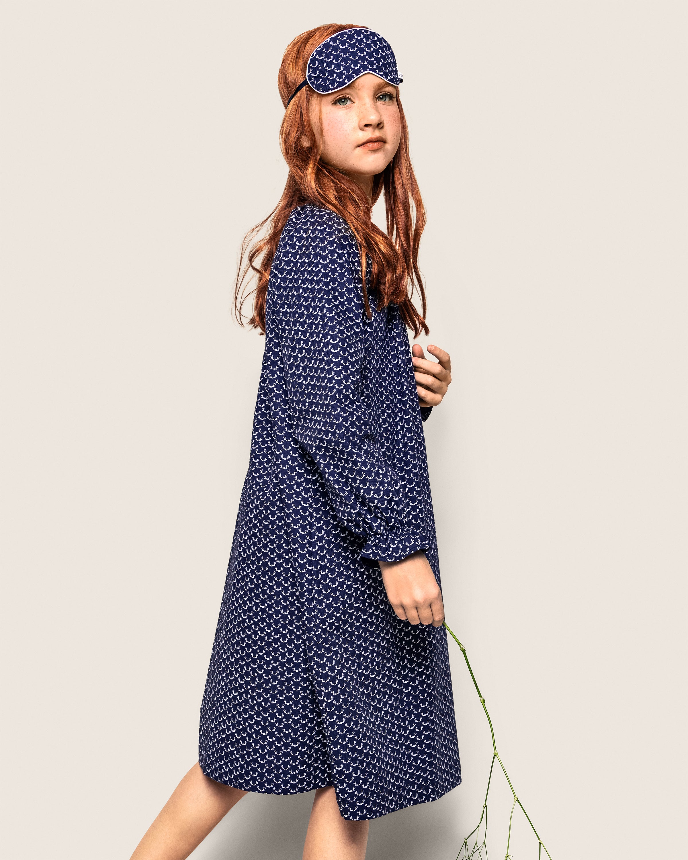 A red-haired girl in Petite Plumes Girls Twill Delphine Nightgown in Nordic Antlers and a matching sleep mask stands against a neutral background, holding a thin branch.