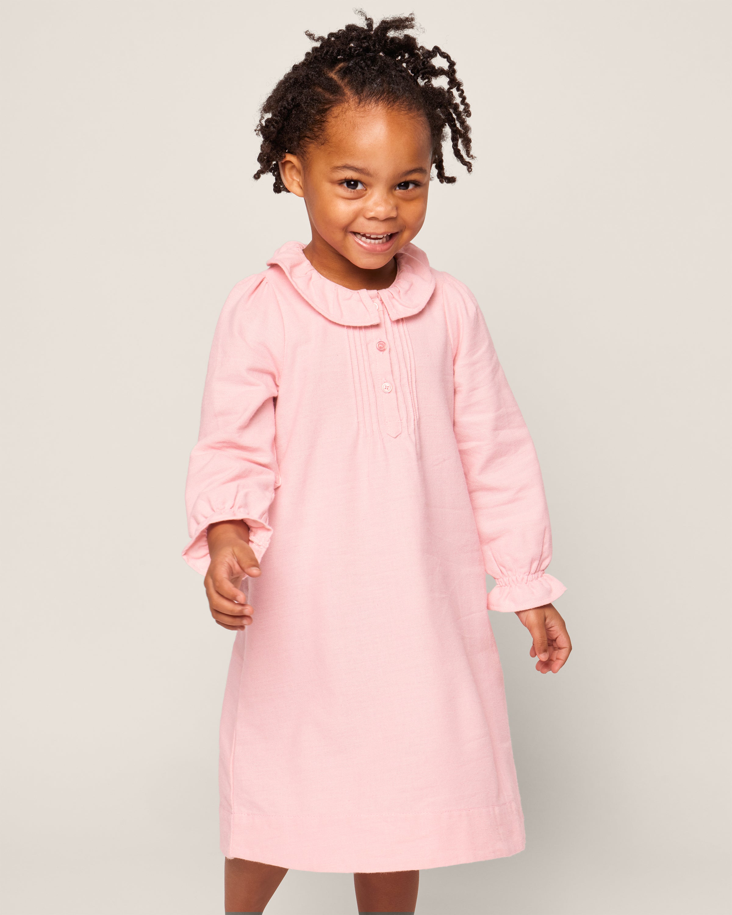 A smiling child with curly hair styled in small twists wears a Petite Plume Girls Flannel Victoria Nightgown in Pink. The backdrop is light beige.