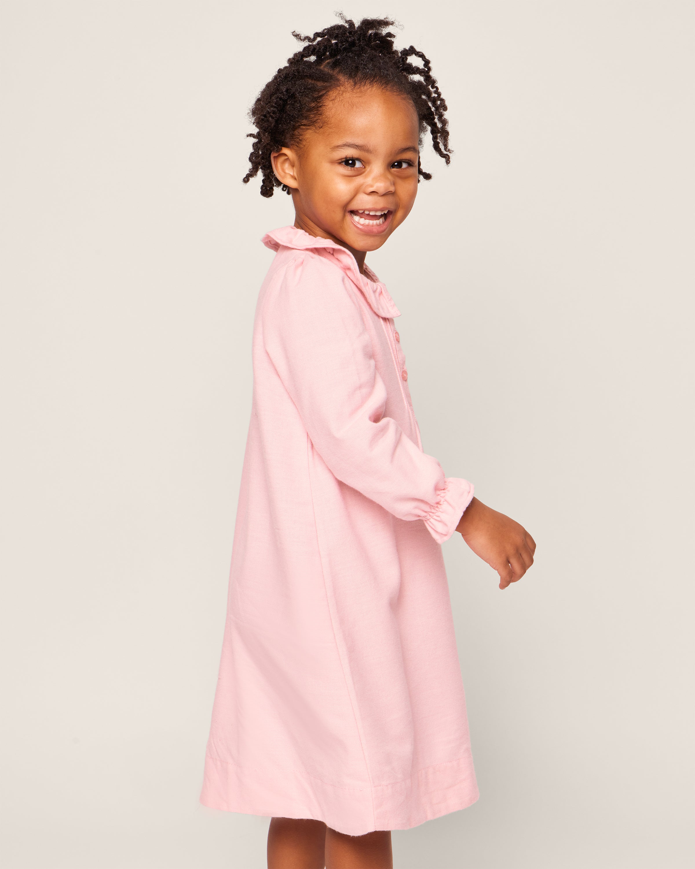 A smiling child with braided hair, wearing a light pink, long-sleeved Girls Flannel Victoria Nightgown by Petite Plume, stands against a plain background. They are turned slightly to the side and looking towards the camera.