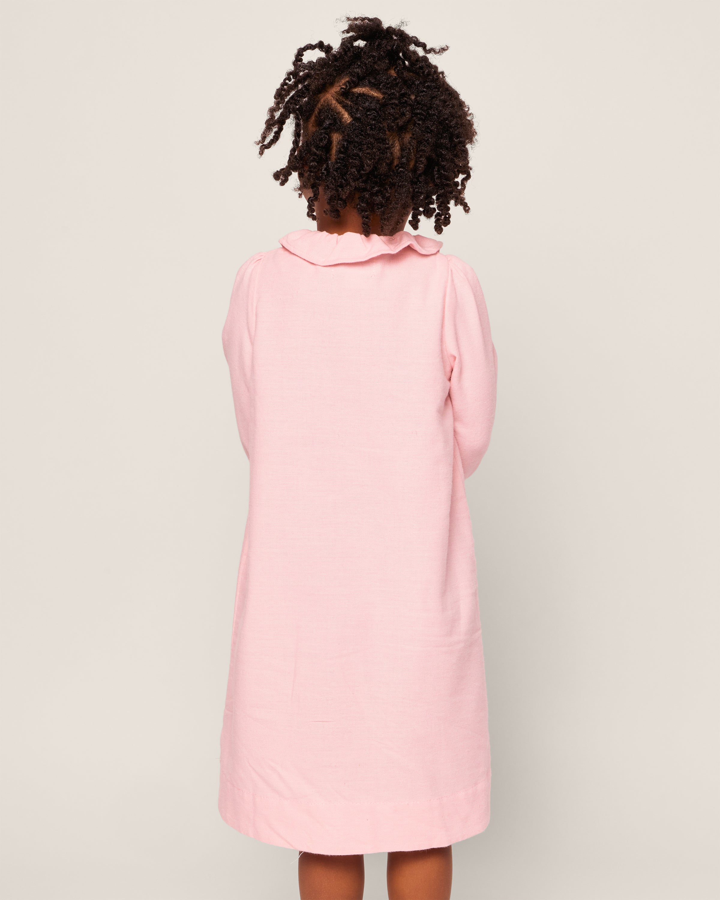 A child is seen from behind wearing a Petite Plume Girls Flannel Victoria Nightgown in Pink, featuring long sleeves and a scalloped collar, against a plain light-colored background.