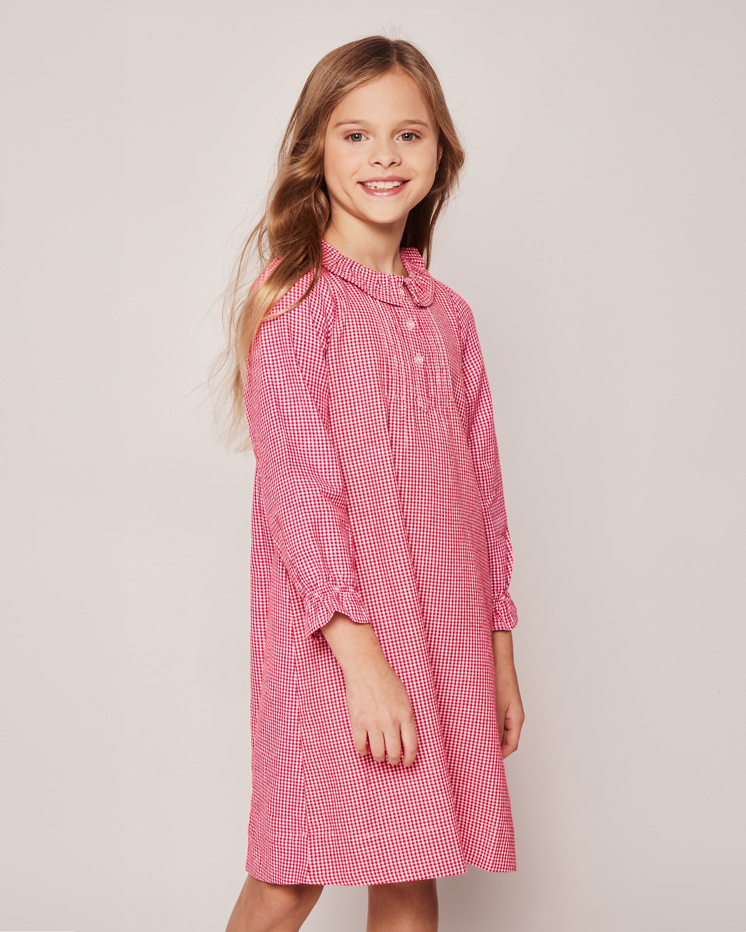 A young girl with long hair, wearing Petite Plumes Girls Flannel Victoria Nightgown in Red Mini Gingham, smiles charmingly against a plain, light-colored background.
