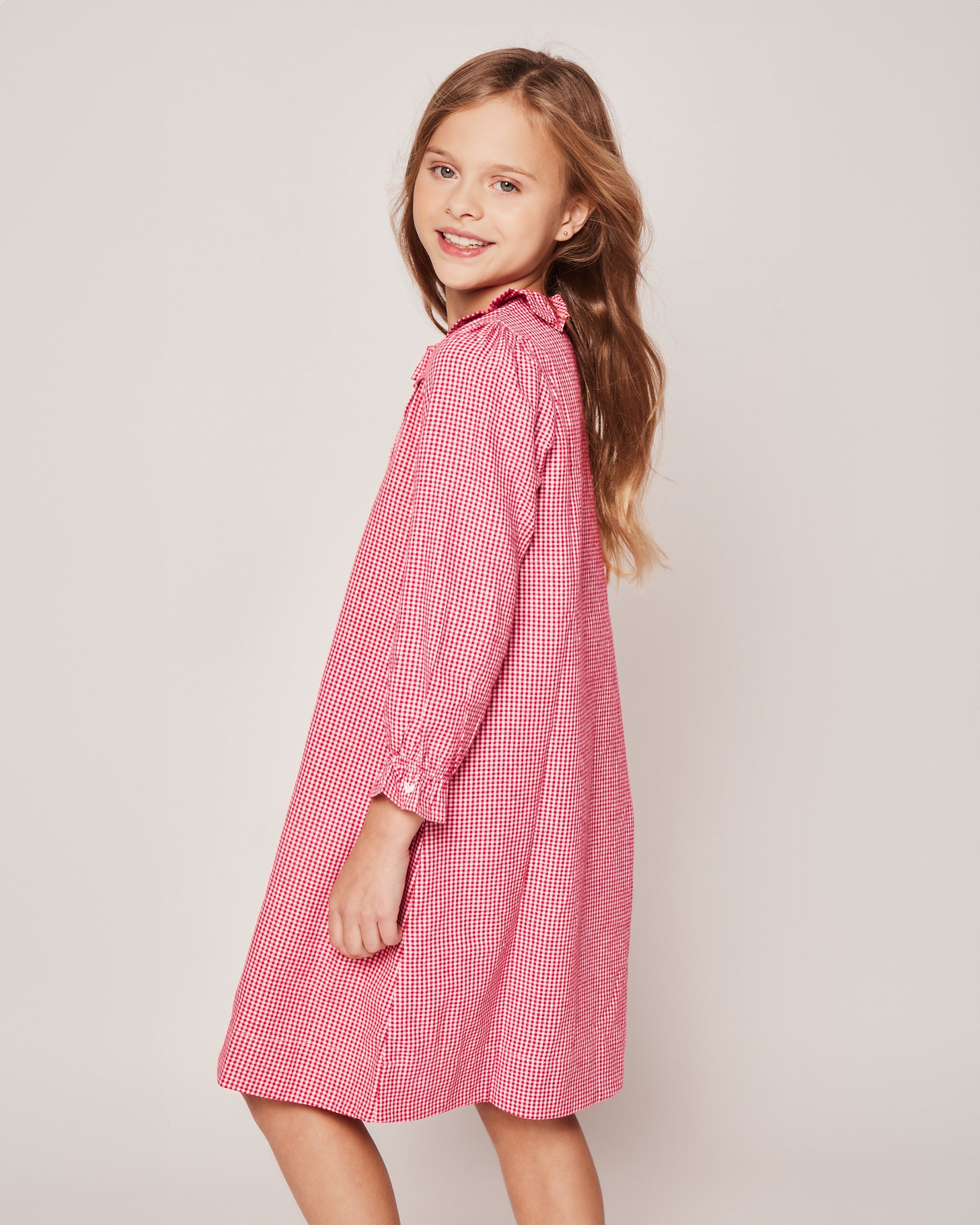 A smiling young girl with long brown hair wears the Petite Plume Girls Flannel Victoria Nightgown in Red Mini Gingham. She stands against a plain background, looking over her shoulder.
