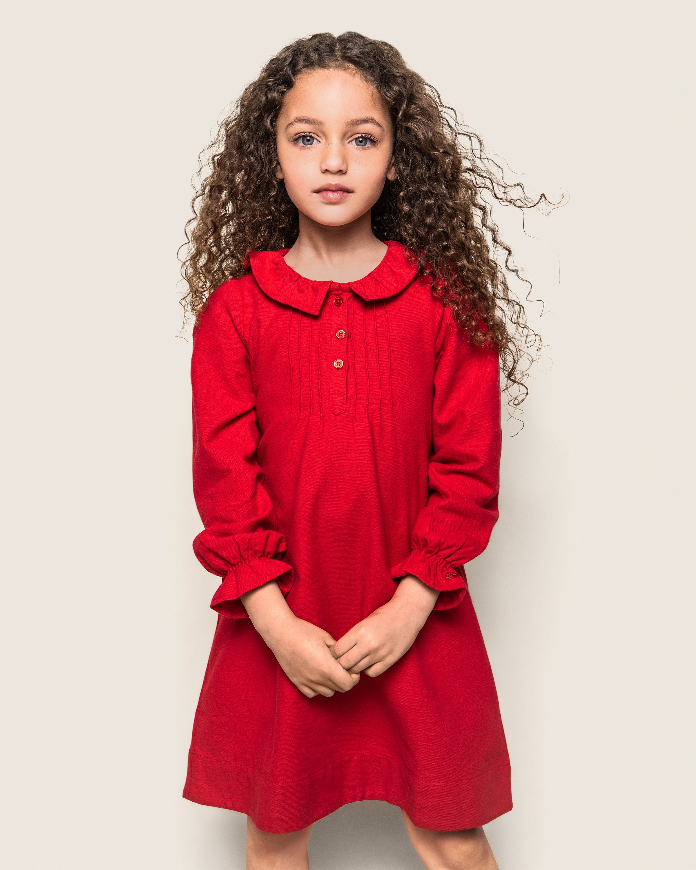 A young girl with long, curly hair stands against a plain background, wearing the Petite Plume Girls Flannel Victoria Nightgown in Red with a collar and buttons. Made from flame-retardant fabric resembling childrens sleepwear, her hands are clasped as she gazes directly at the camera.