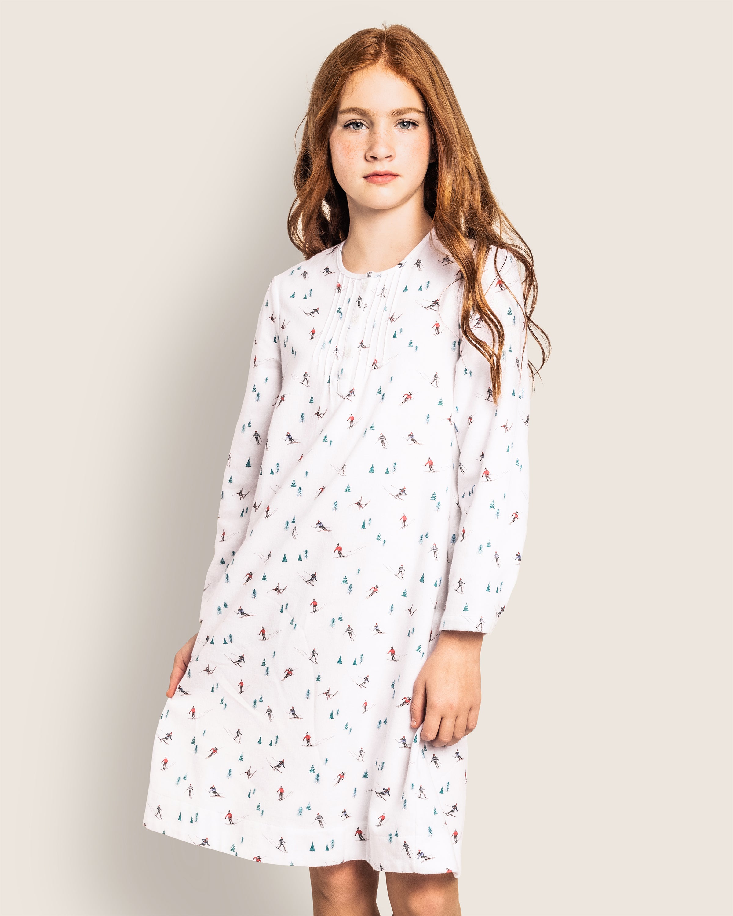 A young girl with long red hair models the Girls Flannel Beatrice Nightgown in Après Ski by Petite Plume. Set against a light beige background, she gazes into the camera with confidence, wearing her colorful-patterned white dress reminiscent of a nightgown.
