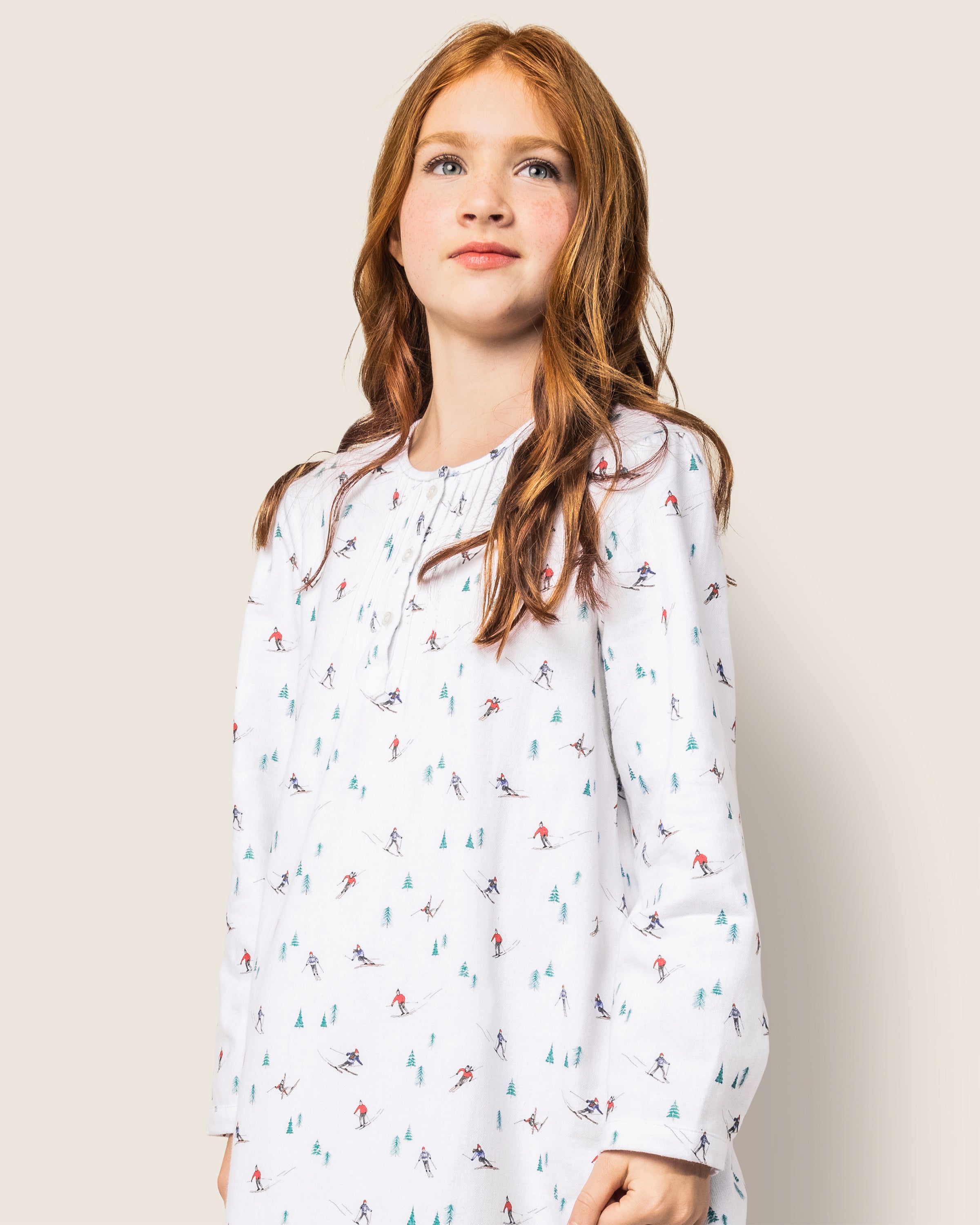A young girl with long red hair wears Petite Plumes Girls Flannel Beatrice Nightgown in Après Ski, featuring a winter-themed skier and trees print. She stands against a plain light background, looking slightly upward.