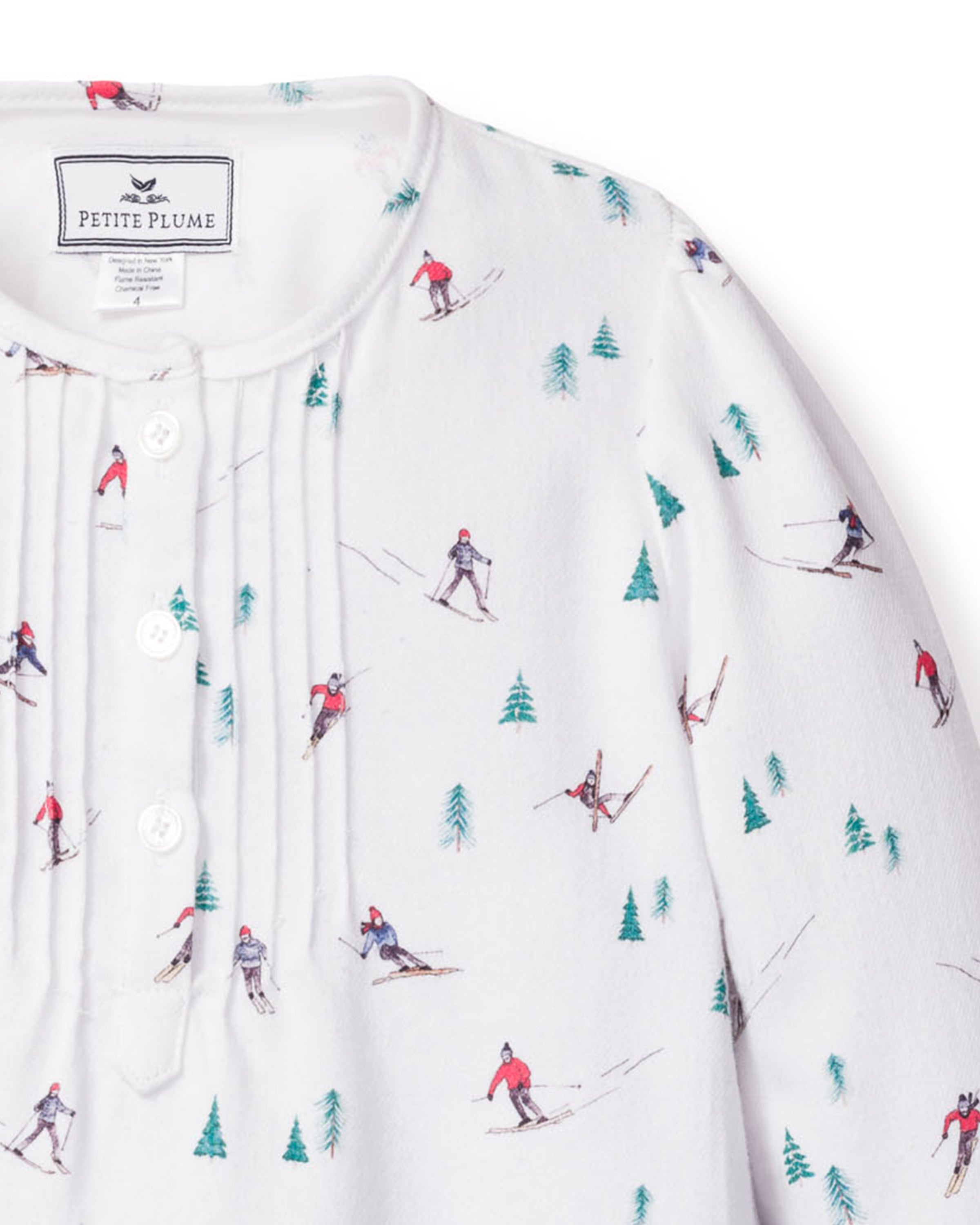 The Girls Flannel Beatrice Nightgown in Après Ski by Petite Plume features white fabric with skiers in red and blue outfits among green pine trees. This luxury cotton piece has a buttoned front, pleats near the neckline, and is ideal for lightweight sleepwear.
