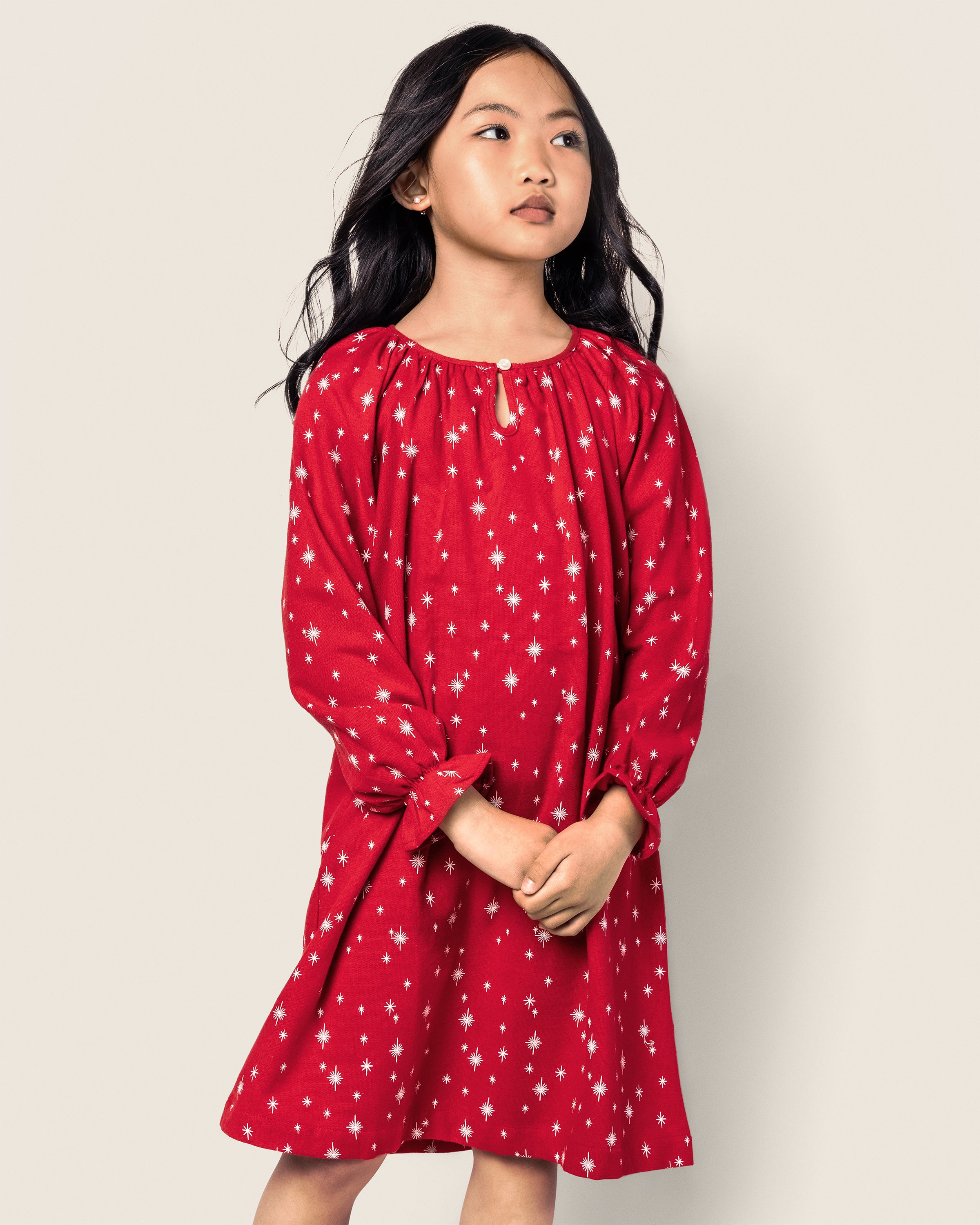 A young girl stands against a plain background, wearing the Girls Flannel Delphine Nightgown in Starry Night by Petite Plume. Her hands are clasped as she gazes slightly upwards, her dark hair cascading over her shoulders in thoughtful repose.