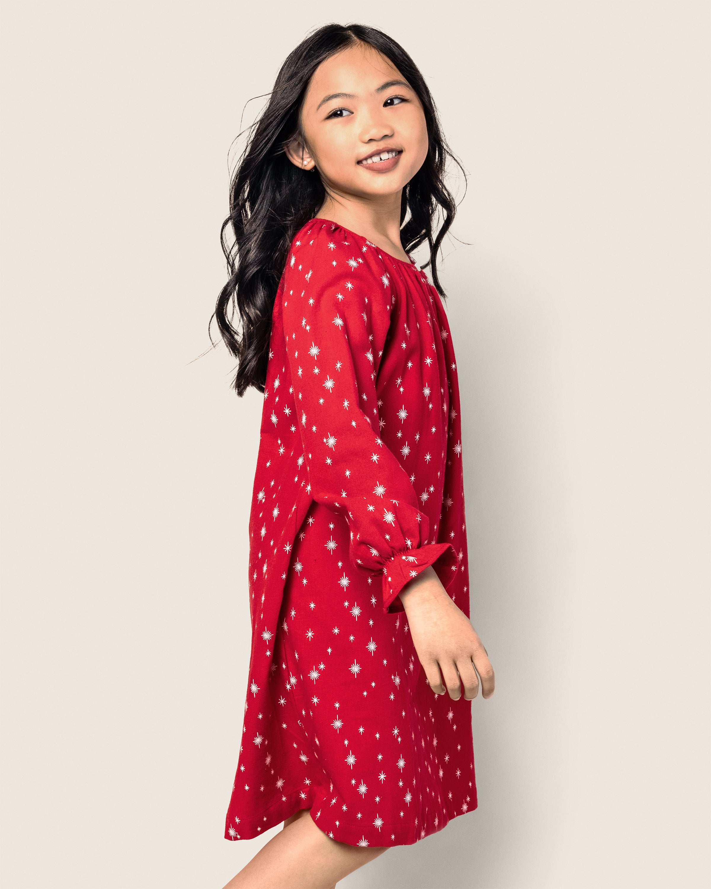 A girl with long dark hair smiles in Petite Plumes Girls Flannel Delphine Nightgown, featuring a red flame-retardant fabric adorned with white star patterns. She turns slightly, looking over her shoulder against a light beige background.