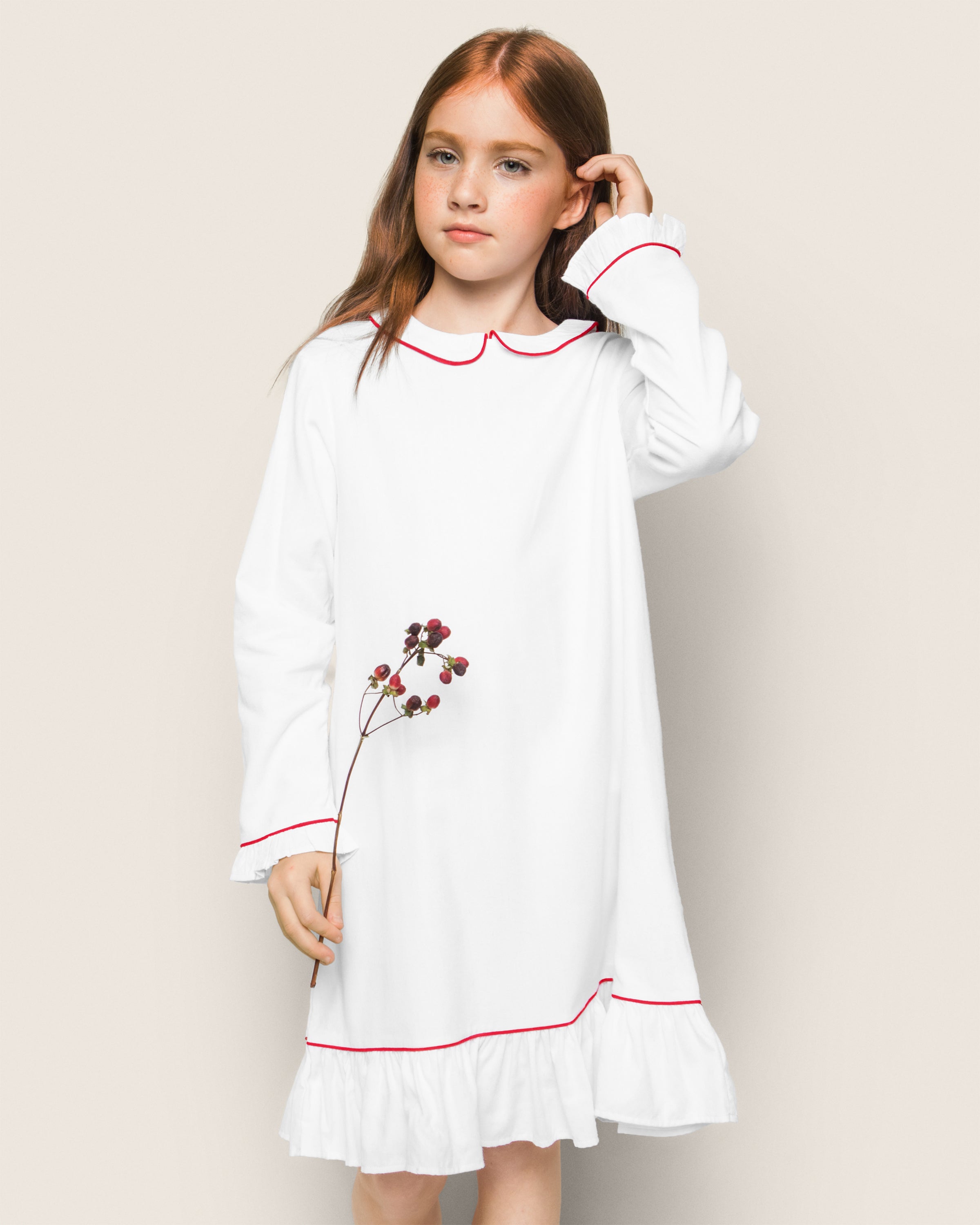A young girl with long red hair wears Petite Plumes Girls White Sophia Nightgown with Red Piping, holding small berries. She stands against a plain light grey background, looking to the side, evoking the cozy comfort of cotton sleepwear.