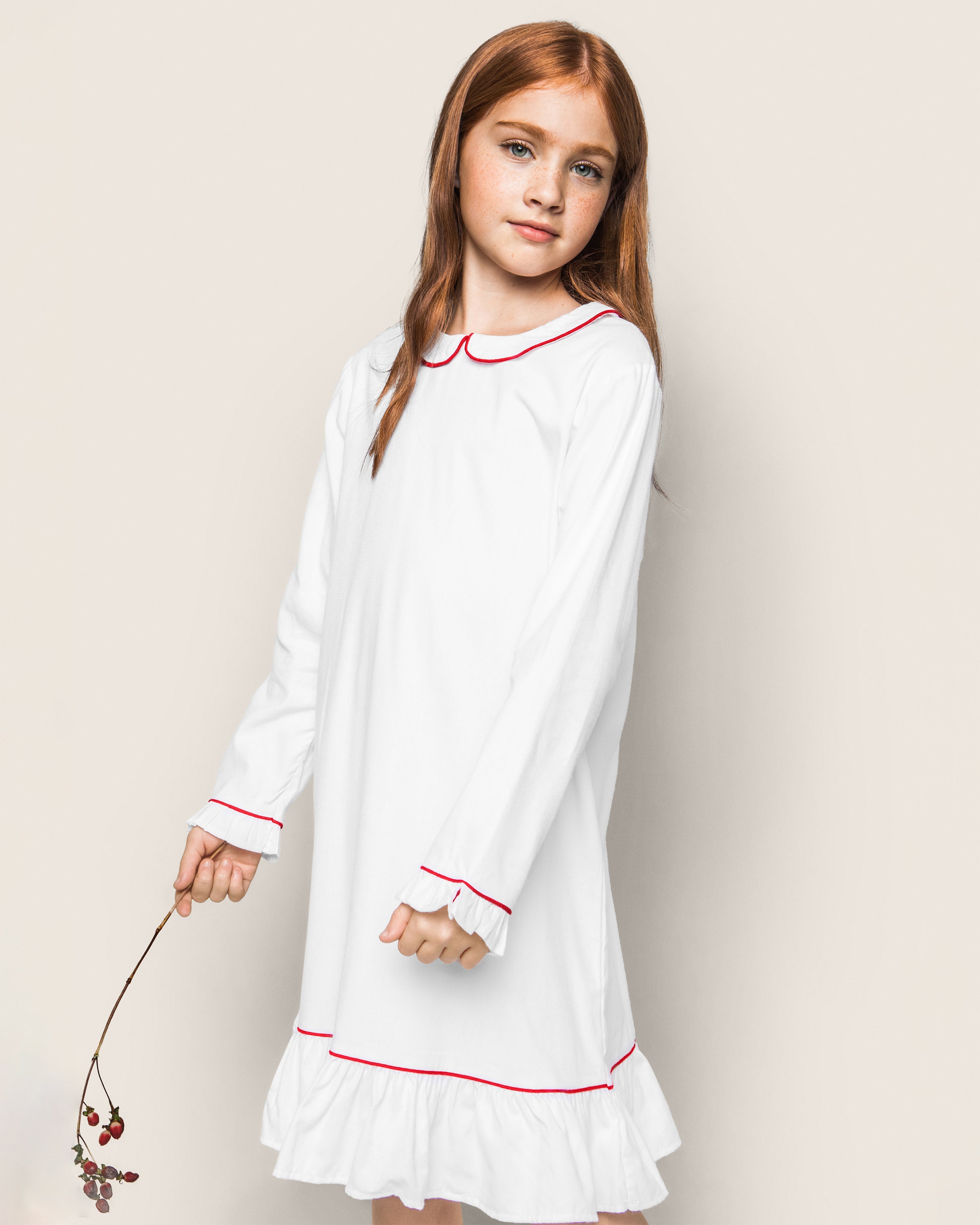 A young girl with long red hair elegantly stands in a Petite Plume Girls White Sophia Nightgown with Red Piping, holding a small branch with red berries against a light gray background, crafting a serene and enchanting scene.