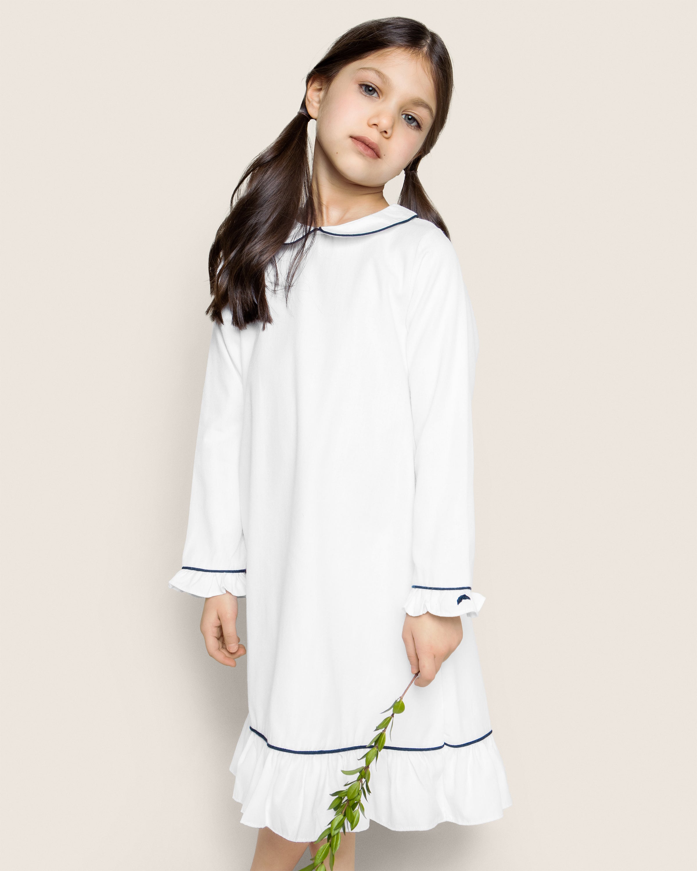A girl with long dark hair in Petite Plumes Girls Twill Sophia Nightgown, featuring navy piping, stands serenely holding a green plant.