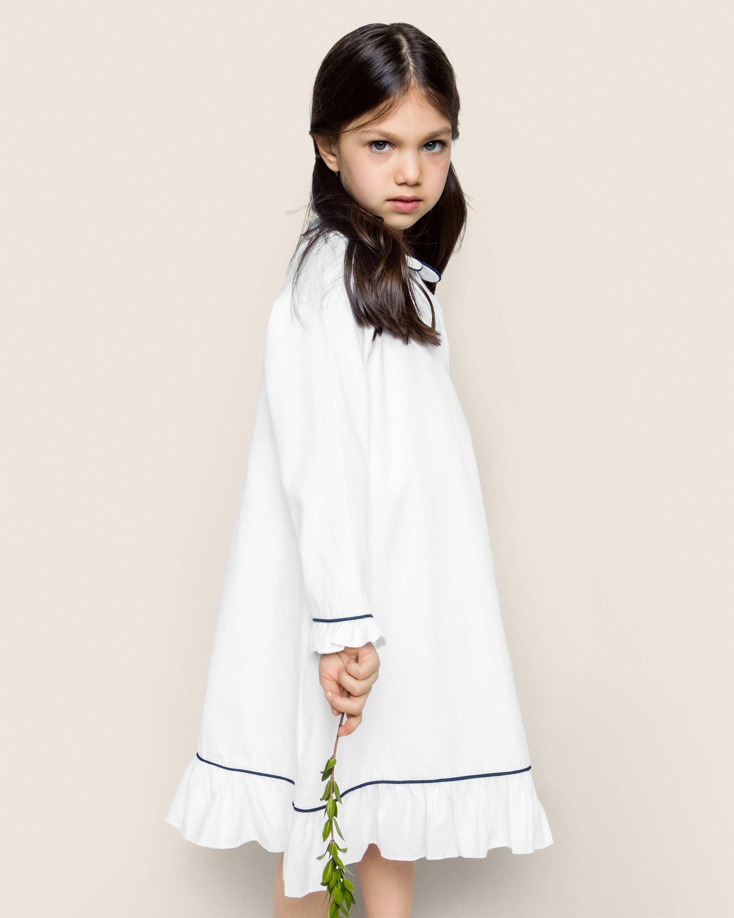 Against a simple backdrop, a young girl with long dark hair wears the Petite Plume Girls Twill Sophia Nightgown in White with Navy Piping. She holds a green plant stem seriously as the sleepwear’s elegant trim and frilled hem complete her serene pose.