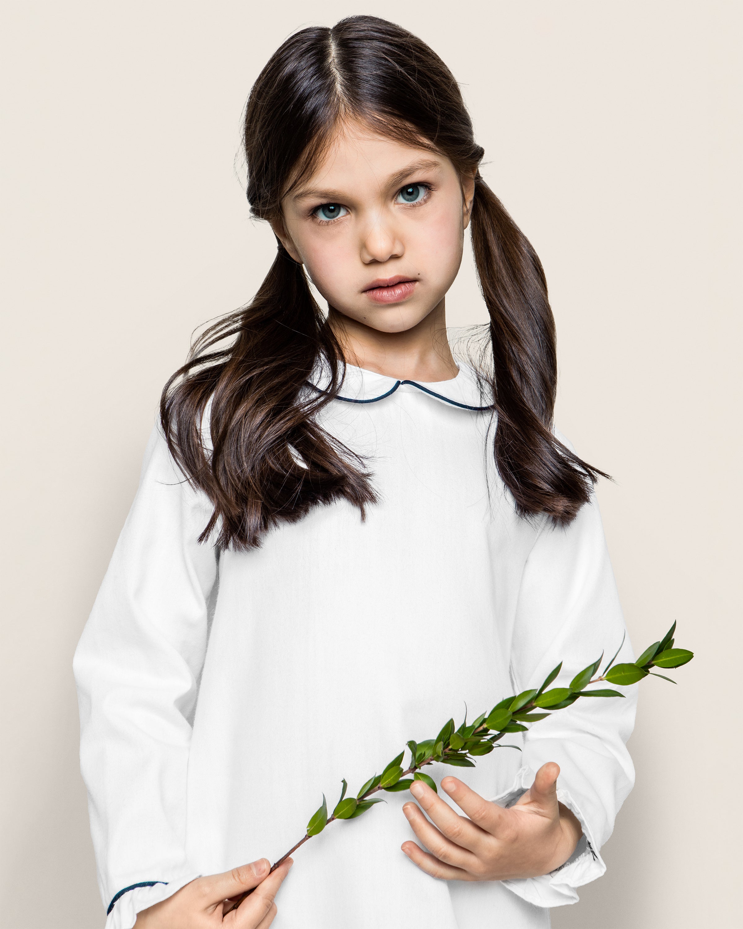 A young girl with long dark hair in pigtails holds a sprig of green leaves, wearing Petite Plumes Girls Twill Sophia Nightgown in White with Navy Piping. Her outfit exudes the classic charm of yarn-dyed cotton as she looks directly at the camera against a neutral background.