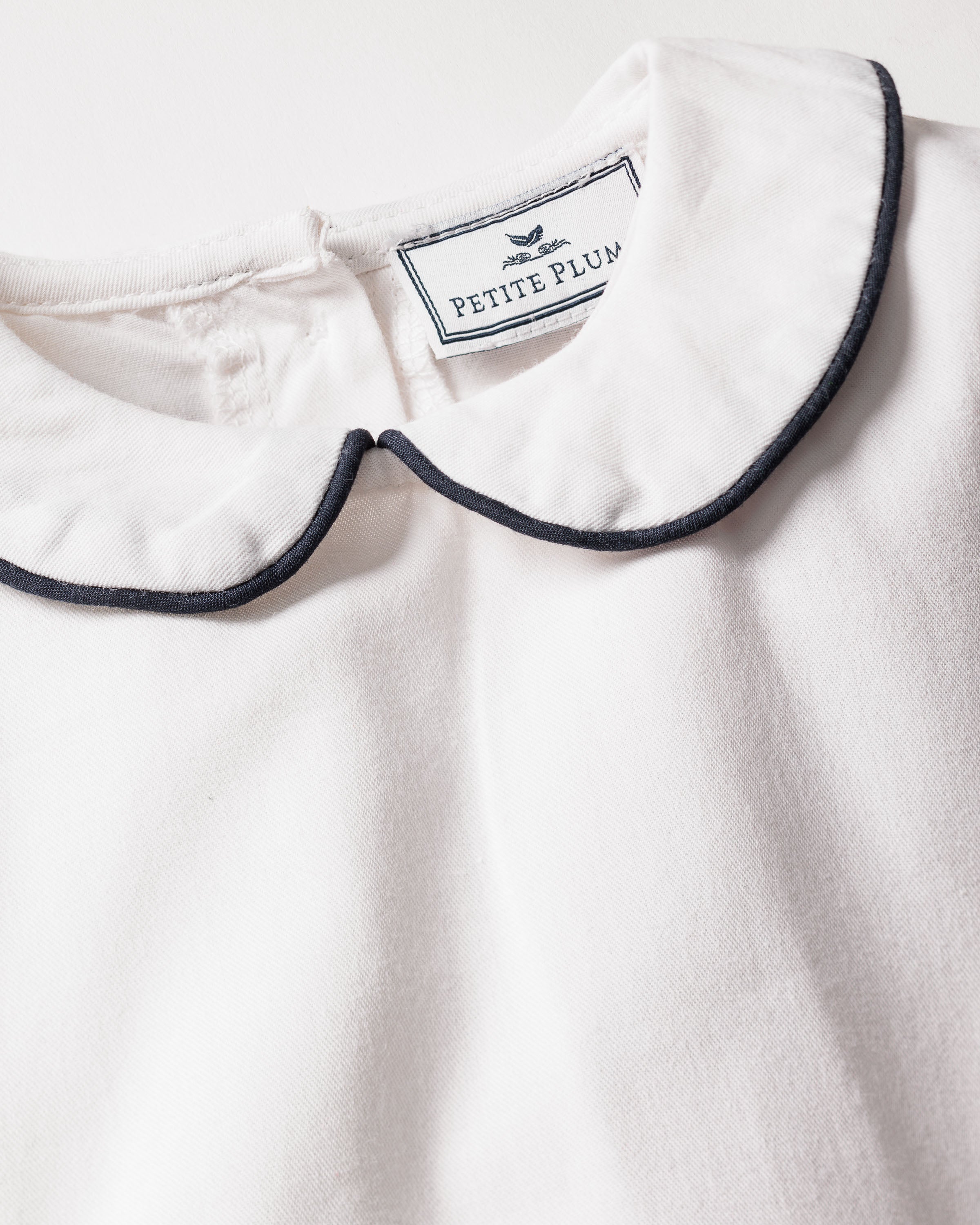 A close-up of the Girls Twill Sophia Nightgown in white with navy piping, featuring a Peter Pan collar and made from yarn-dyed cotton. A neckline label reads Petite Plume, highlighting its luxurious sleepwear origins.