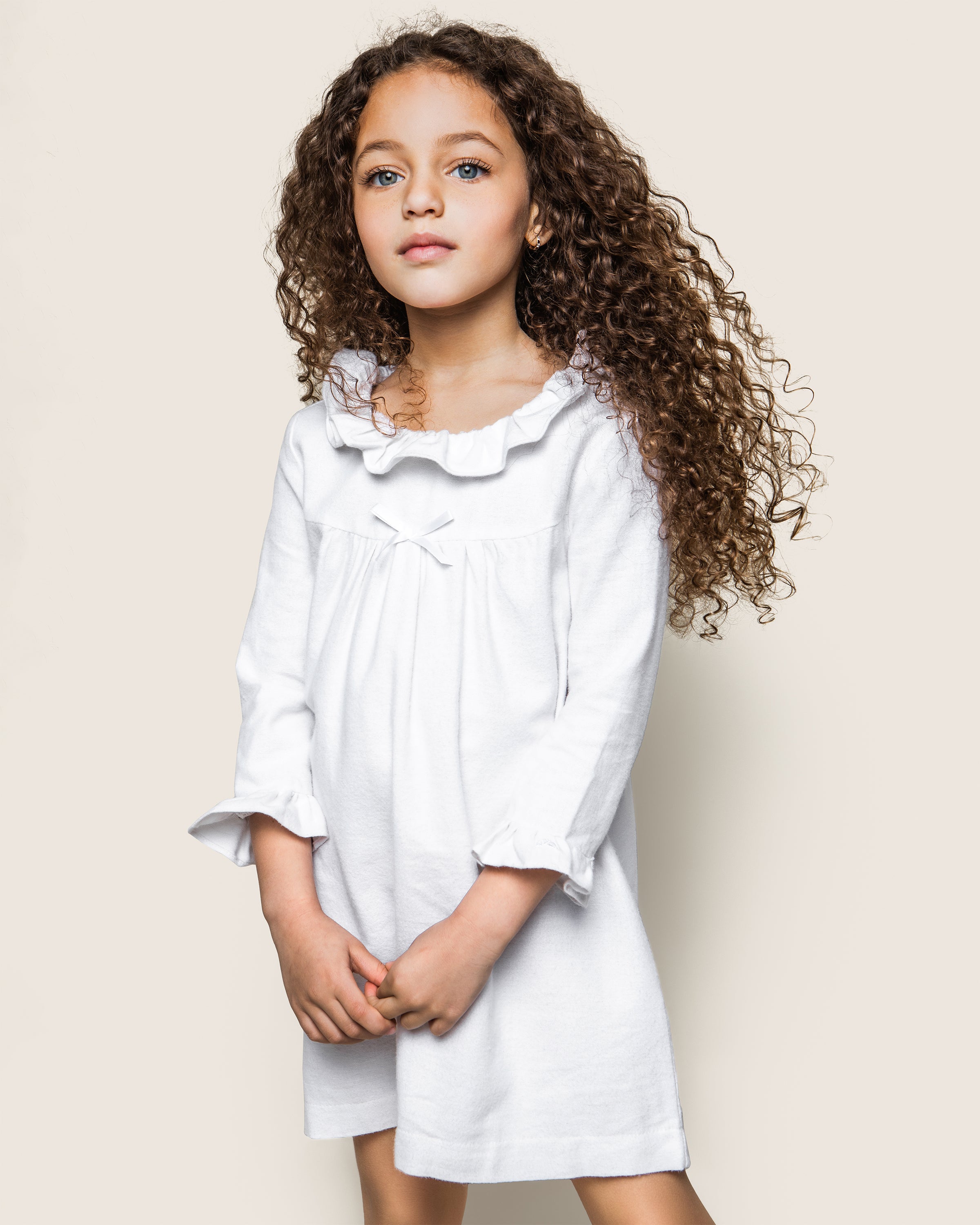 A young girl with long curly brown hair stands against a plain background, wearing the Kids Scarlett Flannel Nightgown in White by Petite Plume. The nightgown features a ruffled collar and sleeves. She gazes directly at the camera with a neutral expression.