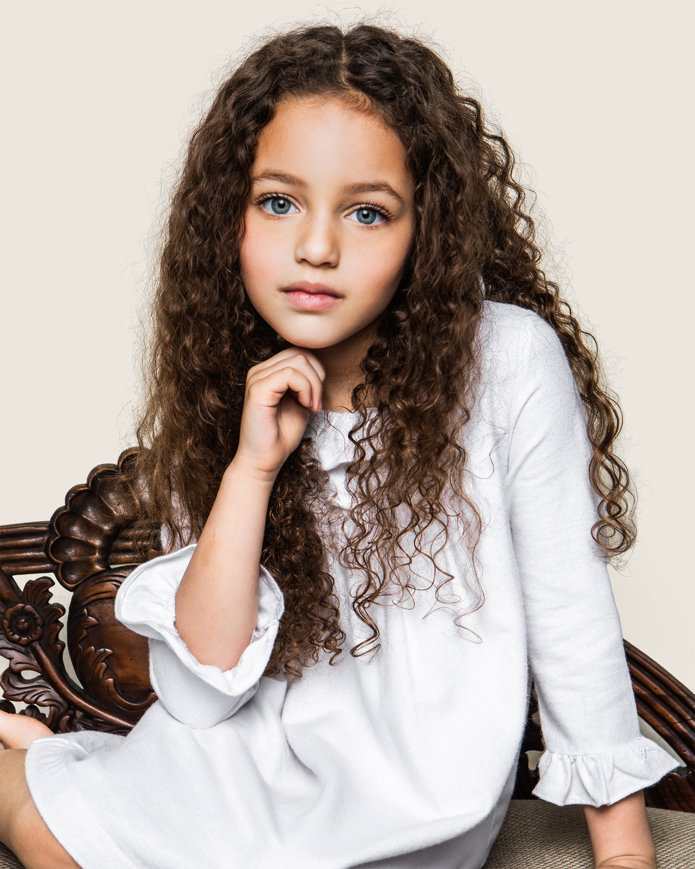 A young girl with flowing curly brown hair and blue eyes wears a Petite Plume Kids Scarlett Flannel Nightgown in white. She sits on a carved wooden chair, chin in hand, looking directly at the camera with a neutral expression against a plain background.