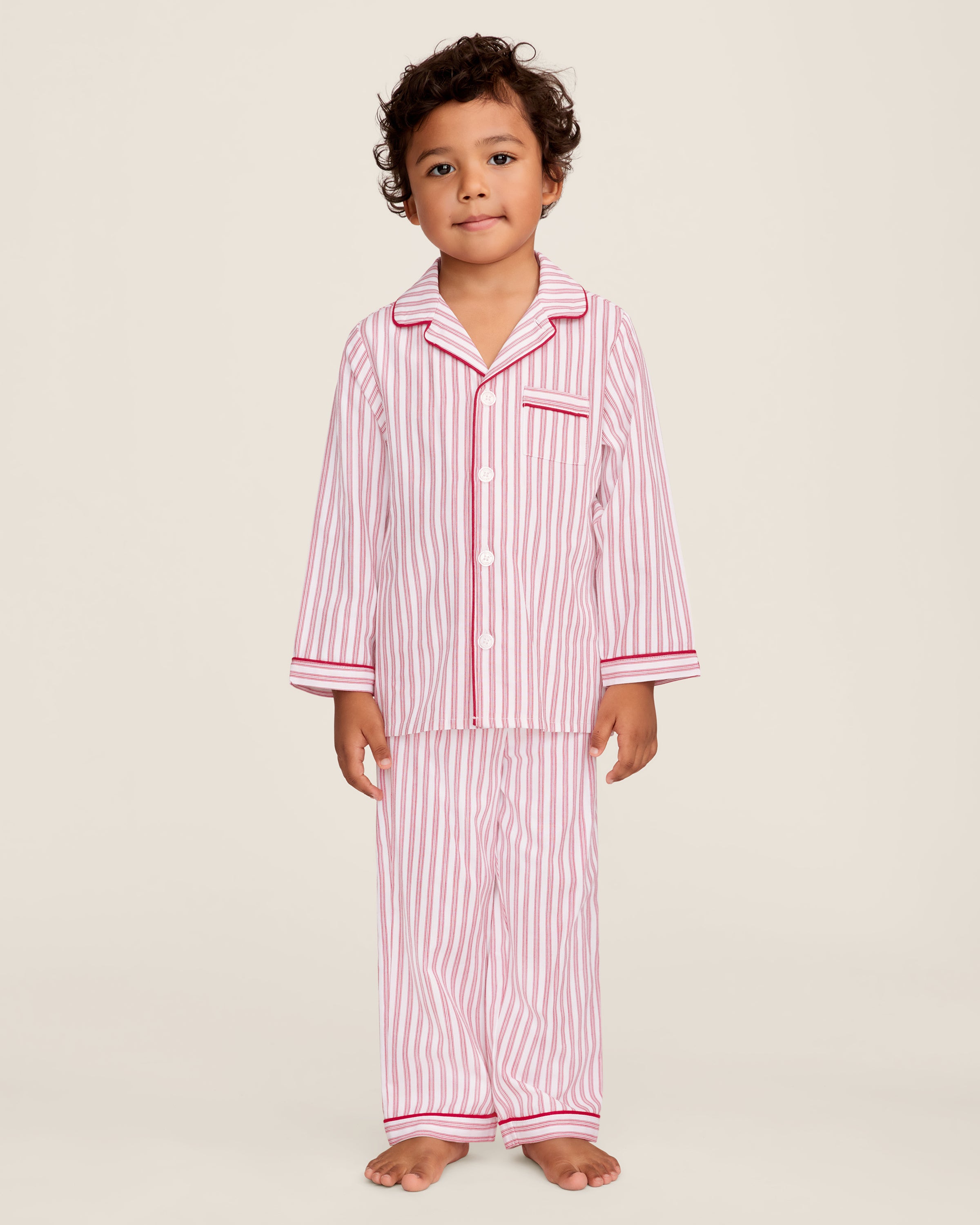 A man and a child sit on a wooden window seat, wearing matching yarn-dyed cotton pajamas. The man wears a pink striped set, while the child dons Petite Plumes Kids Twill Pajama Set in Antique Red Ticking, paired with a red robe. They are reading together.