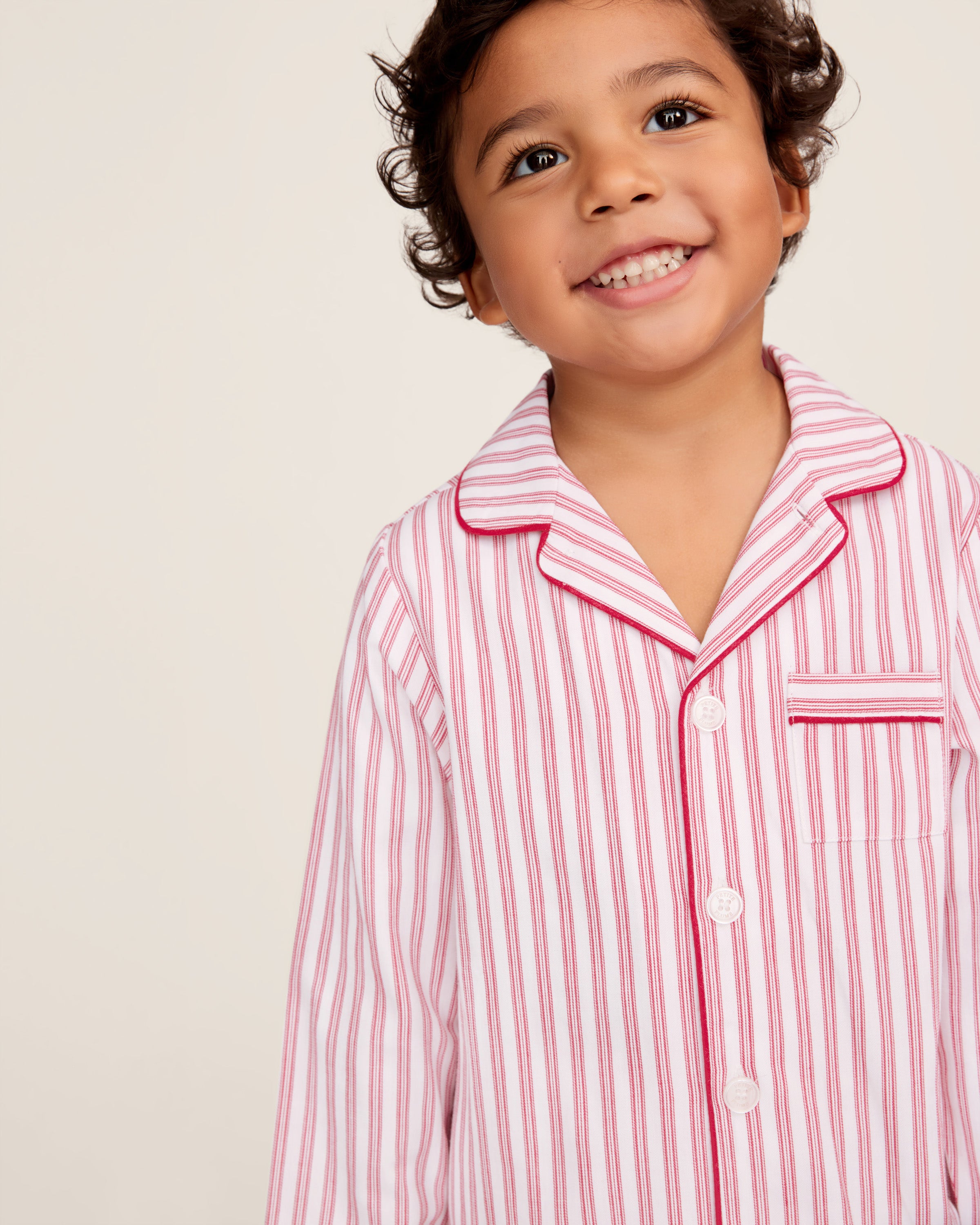 A man and a child sit on a wooden window seat, wearing matching yarn-dyed cotton pajamas. The man wears a pink striped set, while the child dons Petite Plumes Kids Twill Pajama Set in Antique Red Ticking, paired with a red robe. They are reading together.