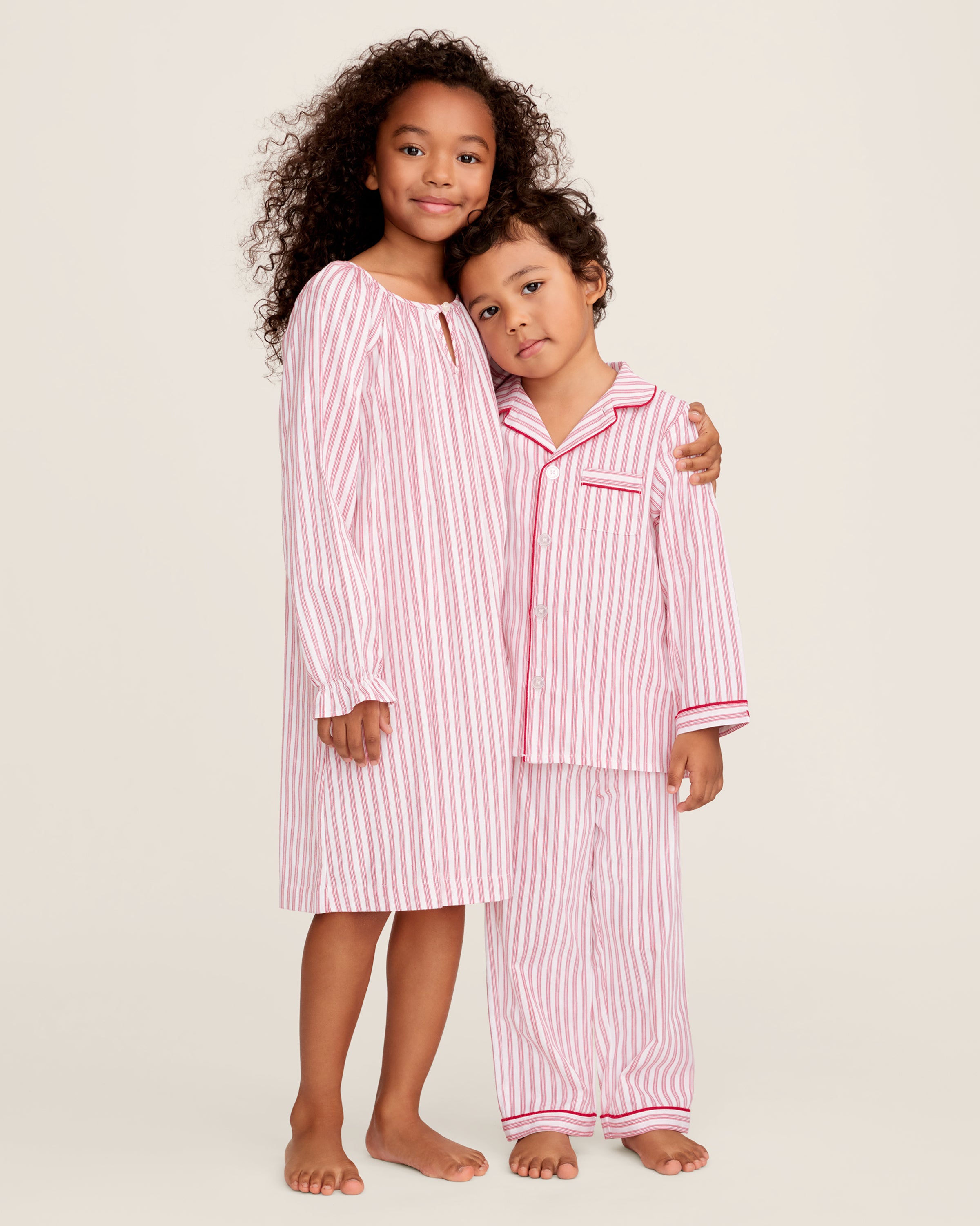 A man and a child sit on a wooden window seat, wearing matching yarn-dyed cotton pajamas. The man wears a pink striped set, while the child dons Petite Plumes Kids Twill Pajama Set in Antique Red Ticking, paired with a red robe. They are reading together.