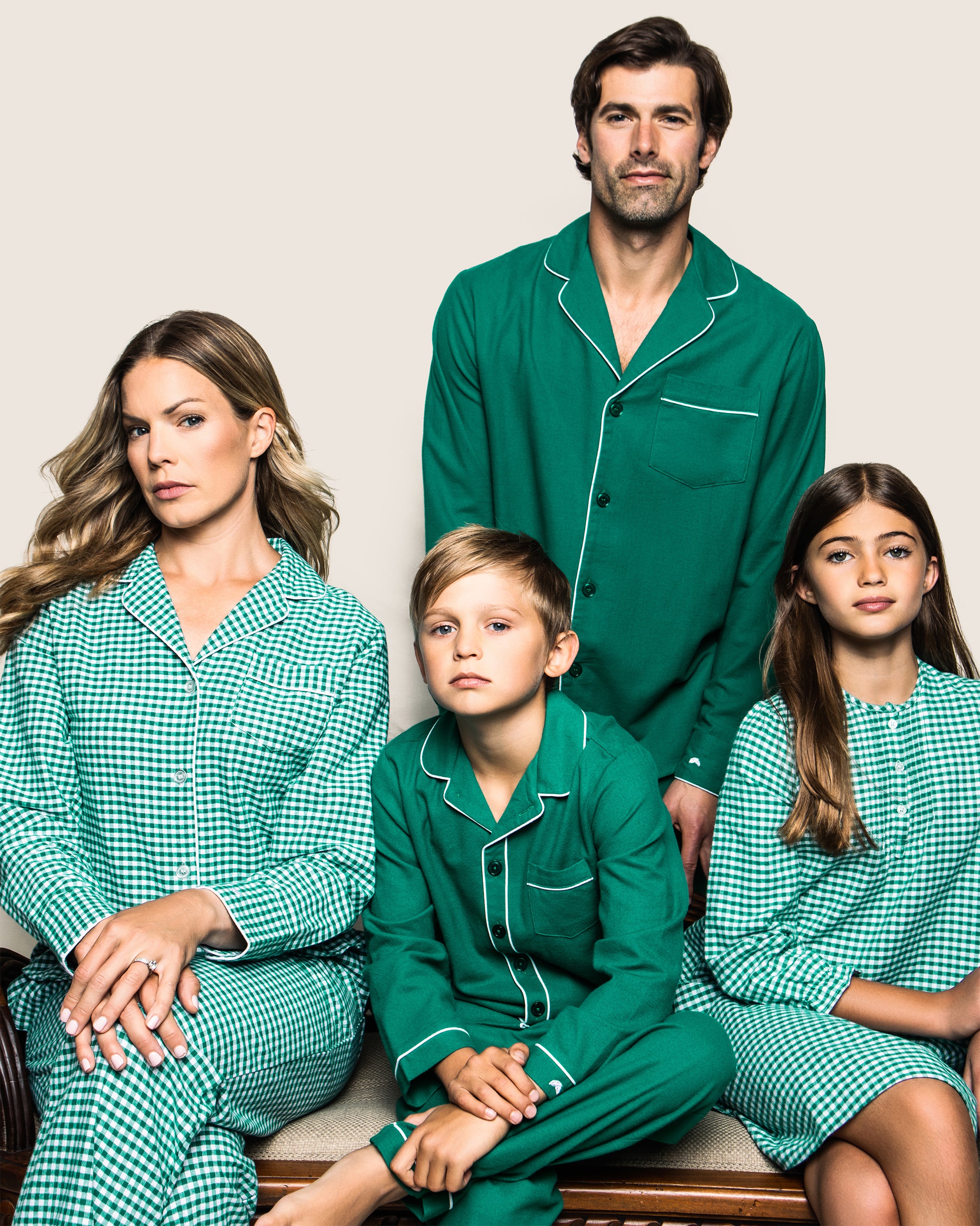 A family of four poses in coordinated sleepwear against a plain background. The adults wear solid and checkered patterns, while the children don Petite Plumes Kids Flannel Pajama Set in Forest Green. Their expressions are serious as they sit closely together.