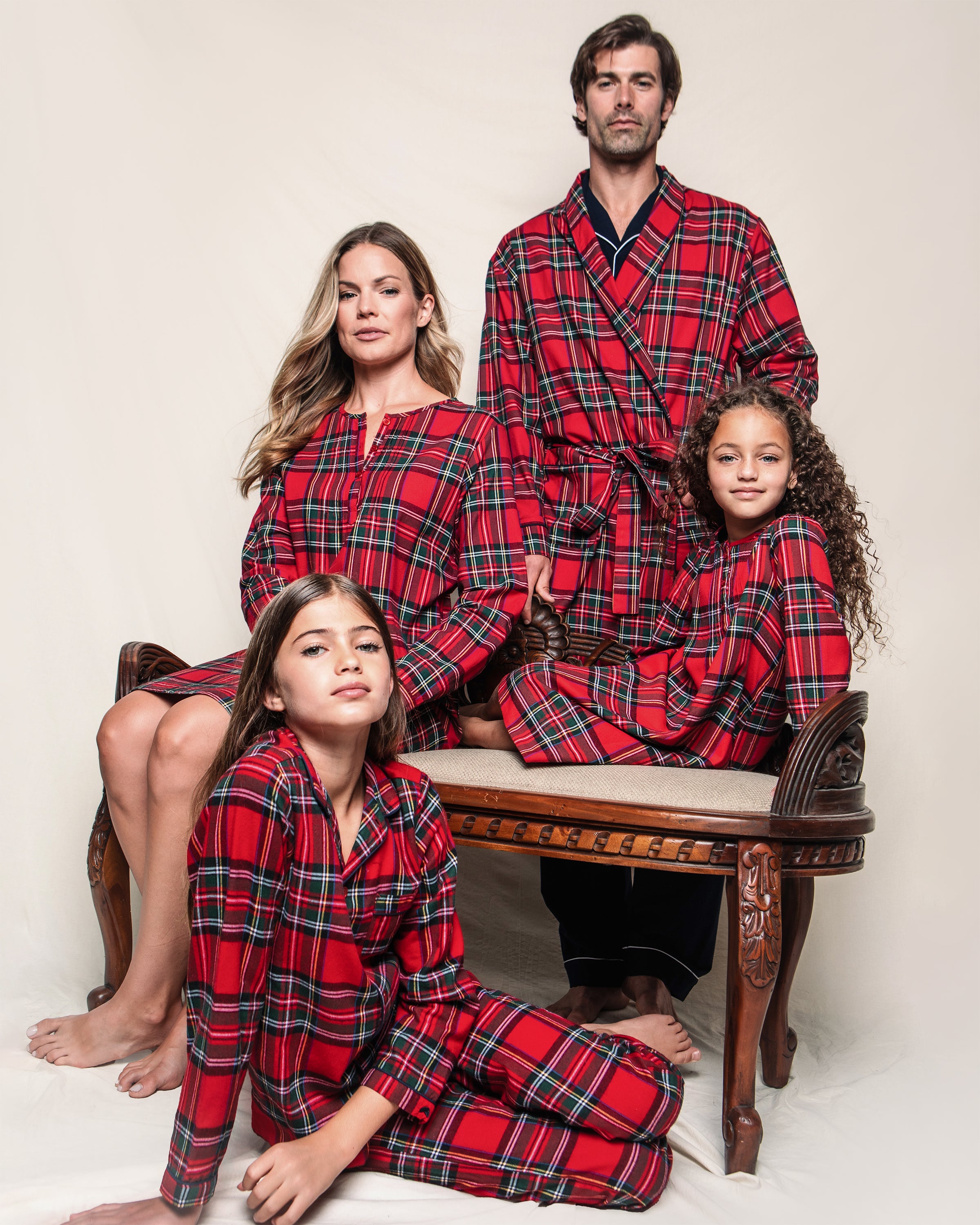 A family of four, dressed in matching Petite Plume Imperial Tartan pajamas, poses barefoot against a neutral background. The parents sit on a bench while their children sit beside them, creating a cozy and coordinated look.