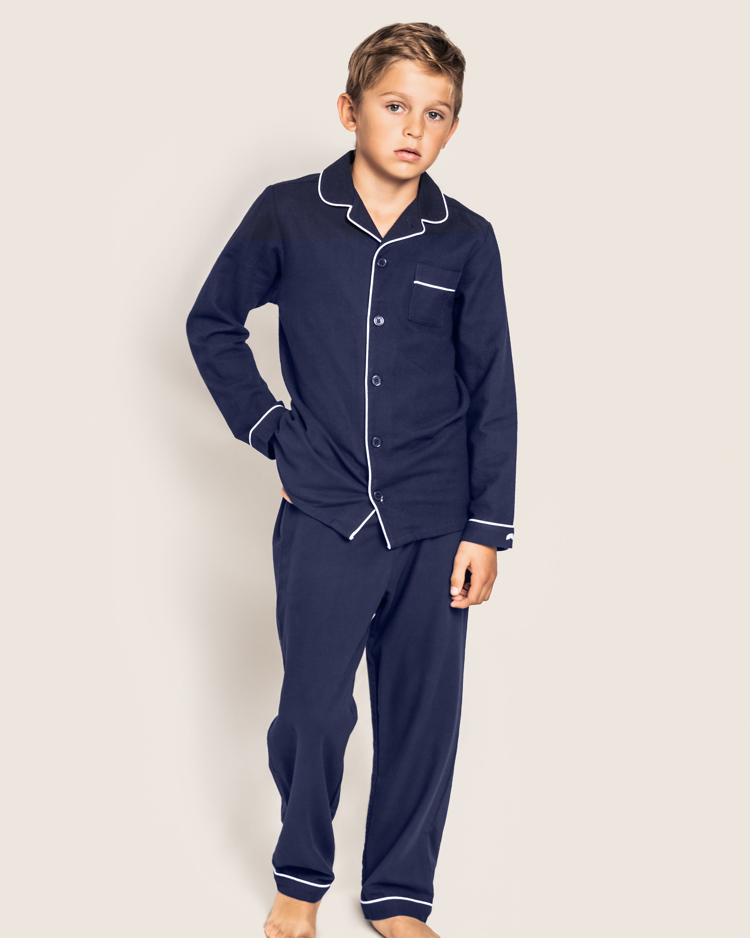 A young boy stands with one hand in his pocket, wearing Petite Plumes Kids Flannel Pajama Set in Navy, featuring white piping and crafted from chemical-free materials. The background is plain and neutral.