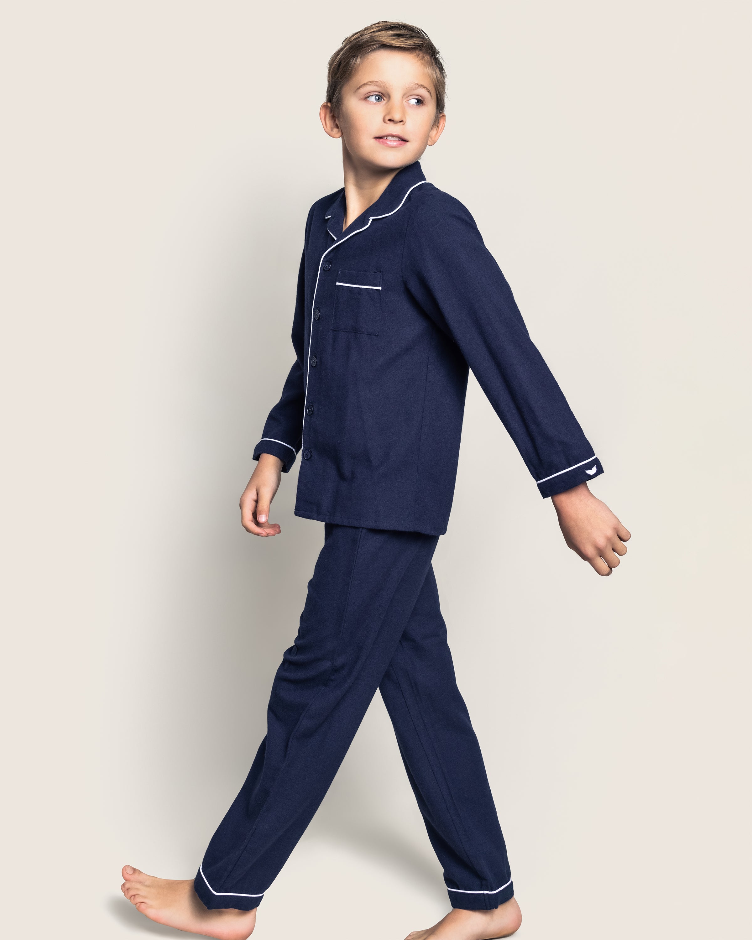 A young boy strolls in Petite Plumes cozy Kids Flannel Pajama Set in Navy, adorned with white piping. Against a plain off-white background, he appears relaxed and casual, his head turned slightly to the side. Crafted from chemical-free sleepwear.