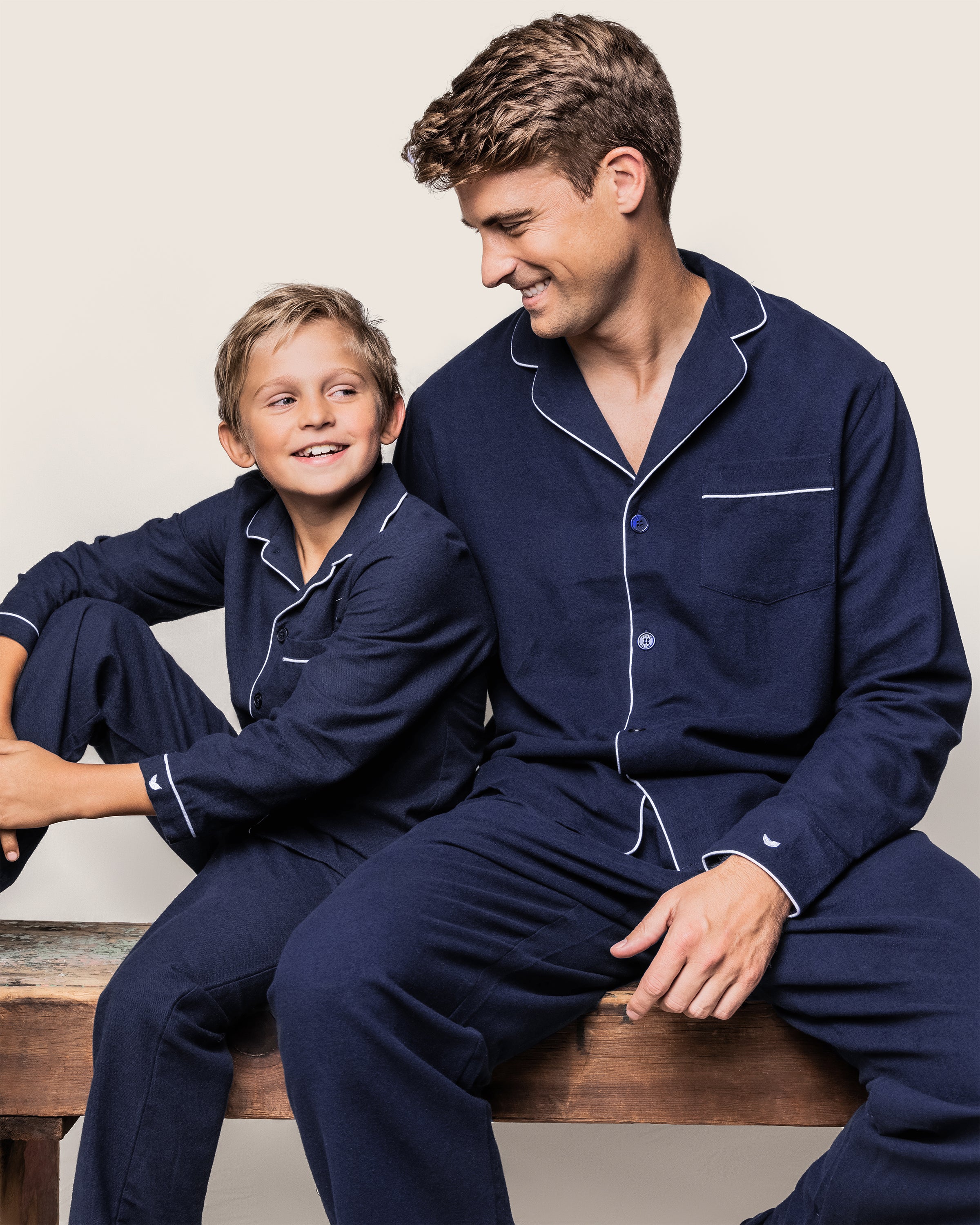 A man and a boy sit on a wooden bench, both smiling at each other against a neutral background. Theyre wearing matching Petite Plumes navy flannel pajama sets, with the boys legs crossed, enjoying their chemical-free sleepwear.