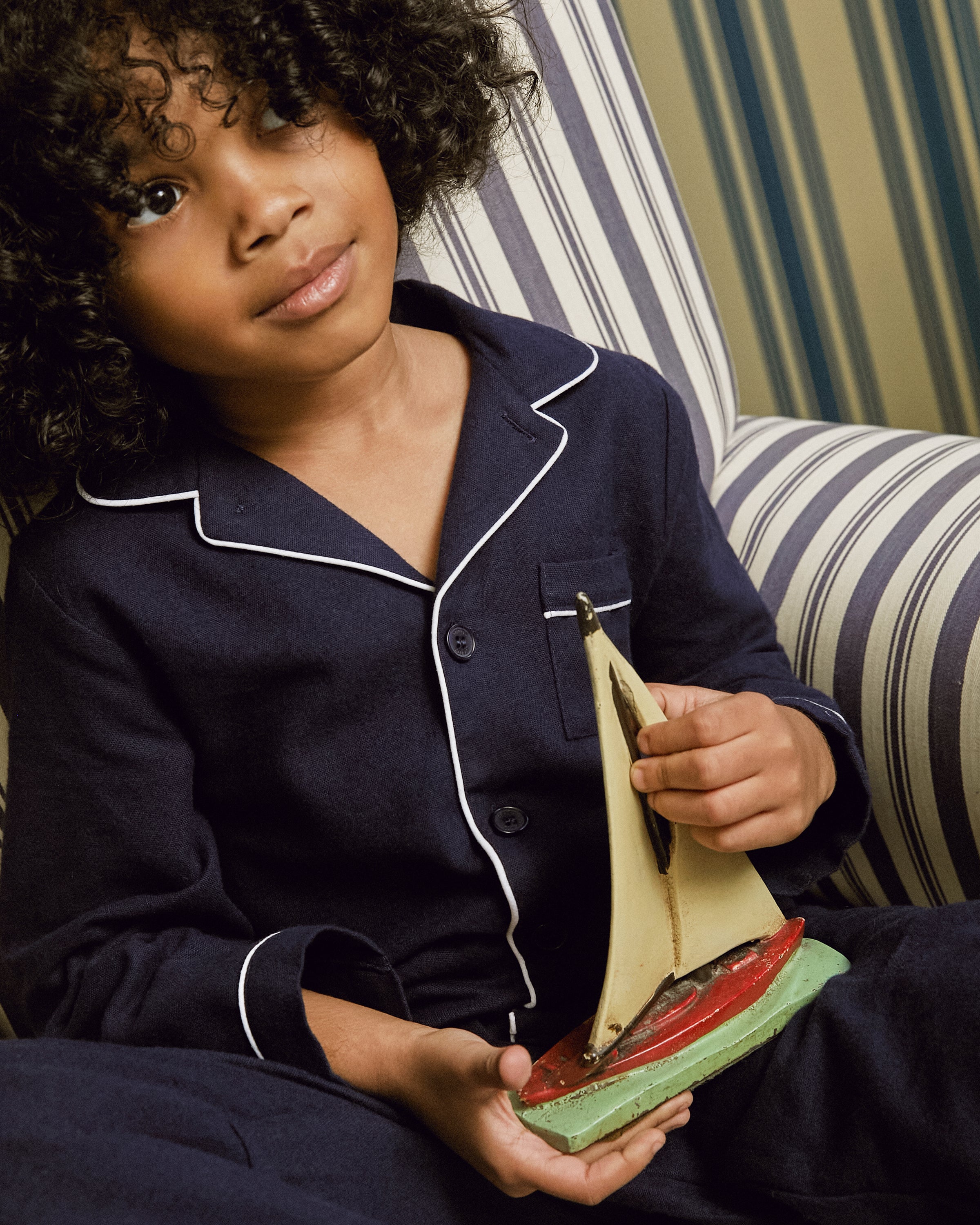 A curly-haired child in Petite Plumes Kids Flannel Pajama Set in Navy, with white trim, sits thoughtfully on a striped armchair holding a model sailboat featuring a red base and cream sail.
