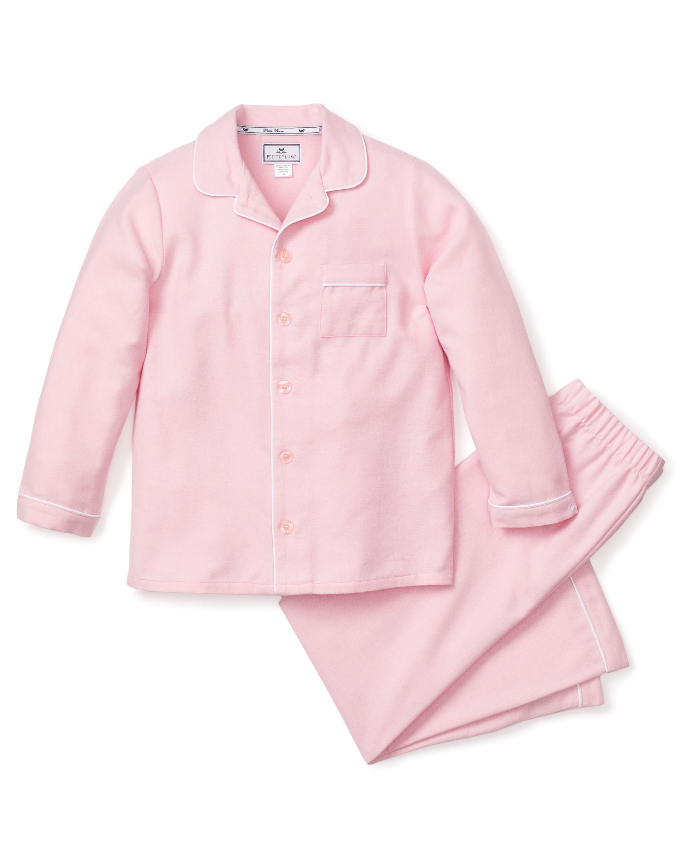The Kids Flannel Pajama Set in Pink by Petite Plume features a luxurious light pink design with white piping, long-sleeved button-up top with a chest pocket, and matching pants with an elastic waistband, displayed on a white background.