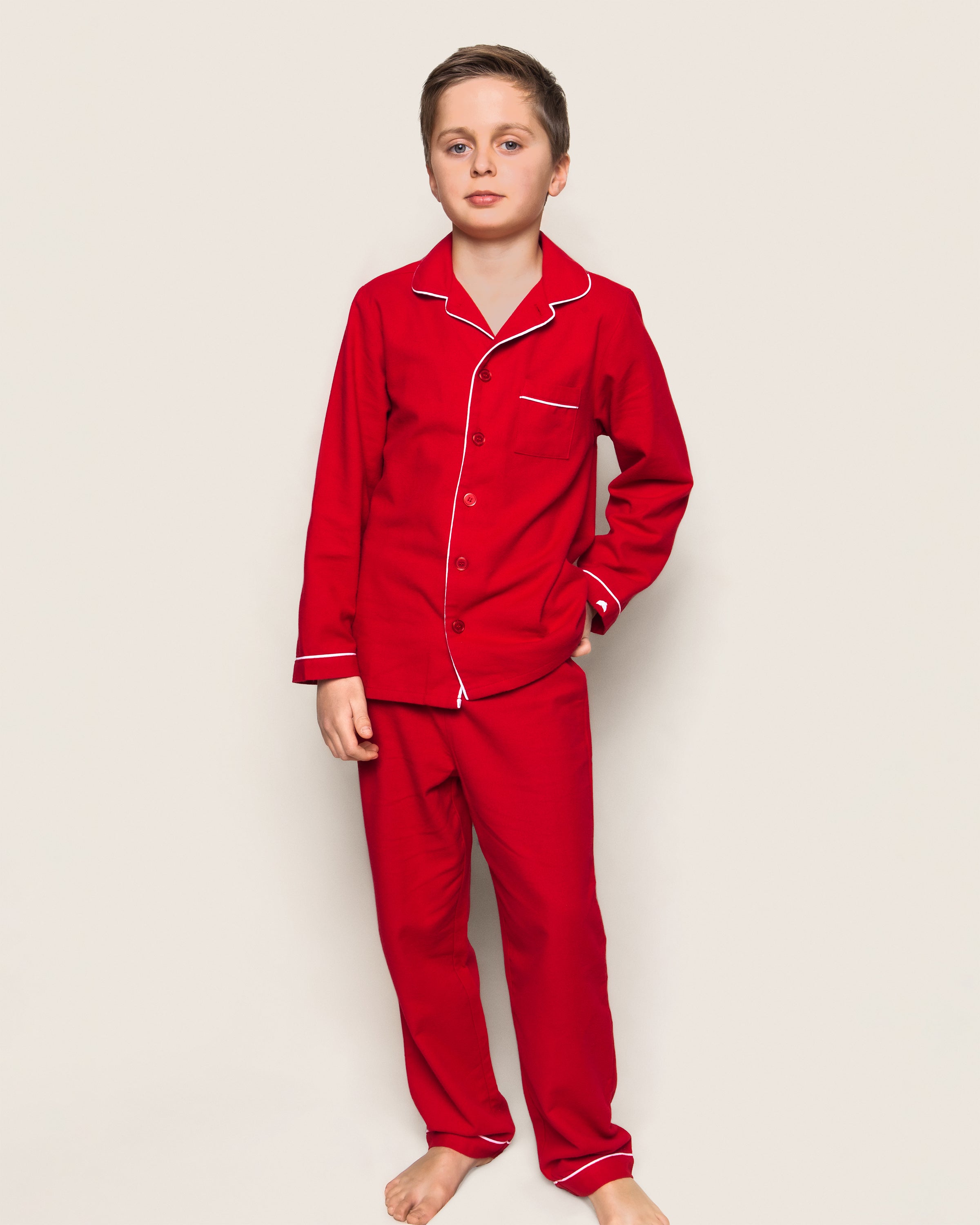 A young boy stands against a light backdrop, modeling Petite Plumes Kids Flannel Pajama Set in Red, featuring cozy white piping. The sleepwear includes a red long-sleeve button-up shirt with matching pants. He is barefoot and appears calm.