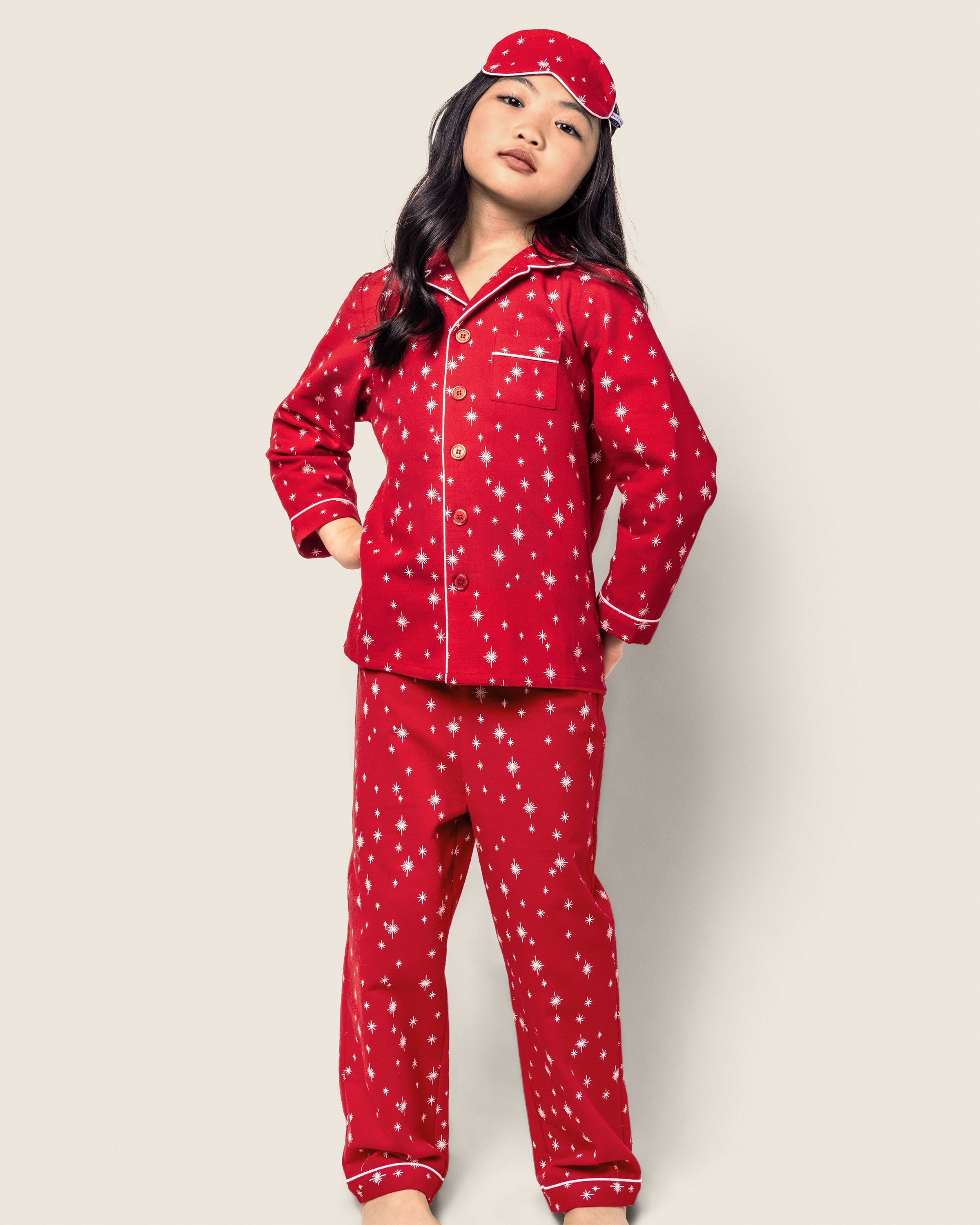 A child stands proudly against a plain backdrop, exuding comfort and style in Petite Plumes Kids Flannel Pajama Set in Starry Night, featuring a red design with white star patterns and a matching sleep mask.