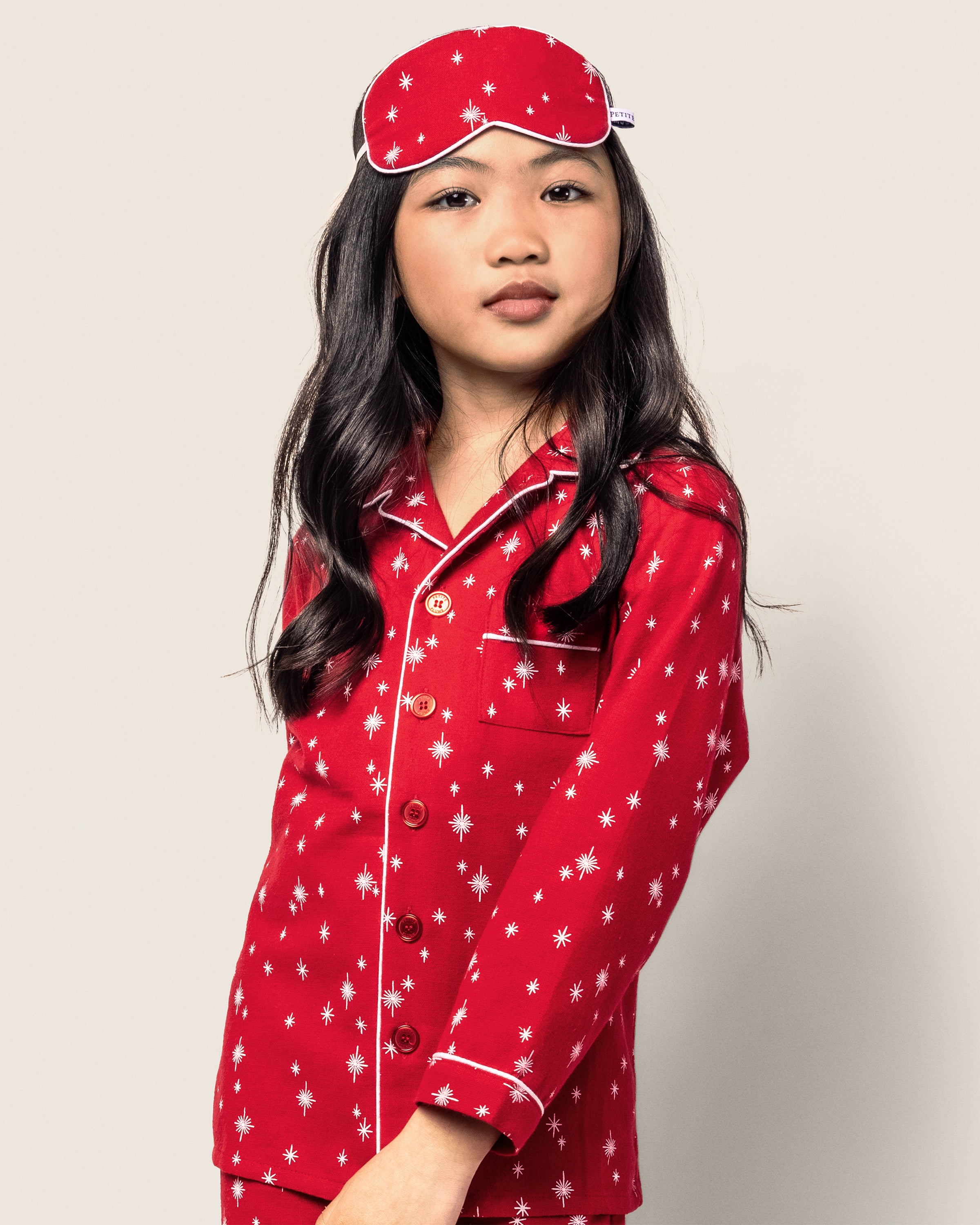 A young girl with long black hair wears Petite Plumes Kids Flannel Pajama Set in Starry Night, featuring a vibrant red star pattern. Made from soft yarn-dyed cotton, the set includes a matching red sleep mask on her head. The background is plain and light-colored.