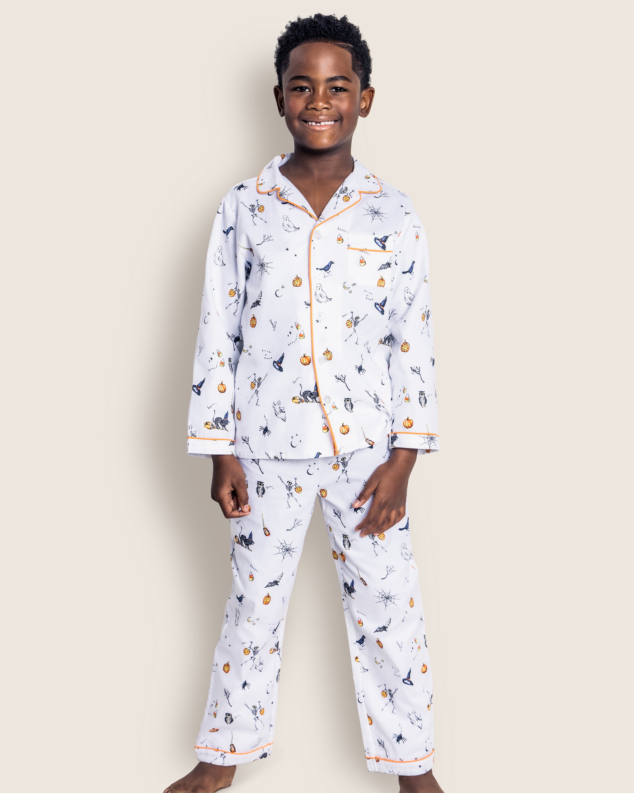 A child grins, dressed in Petite Plumes Kids Twill Pajama Set in Trick or Treat—luxury sleepwear with a colorful creature print and orange trim on the collar, cuffs, and pockets. The backdrop is a plain light beige.